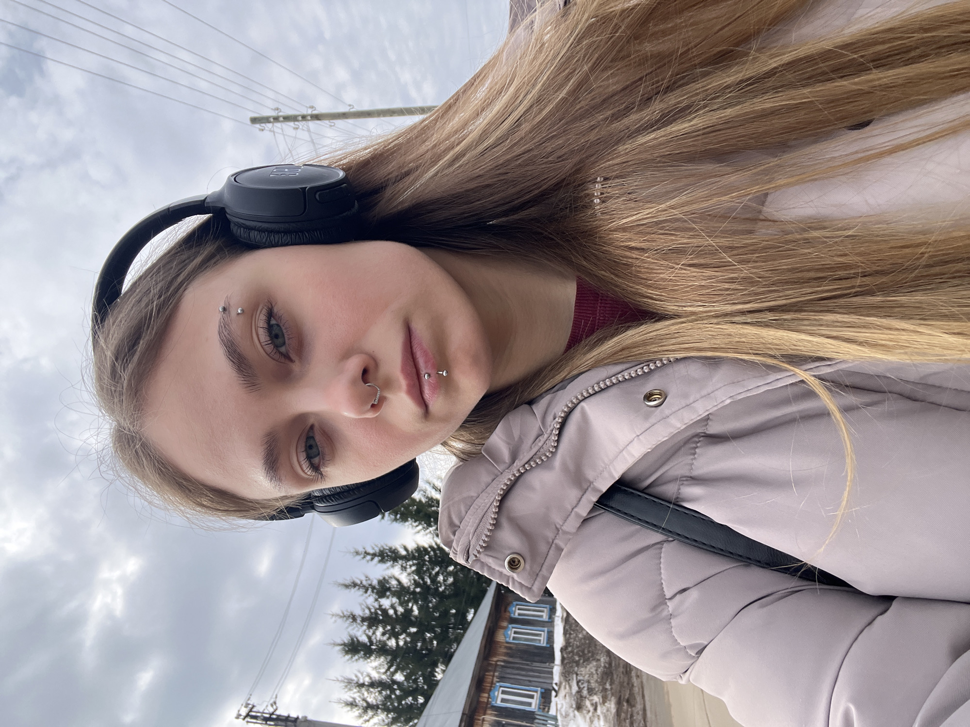 The photograph is a selfie-style image of a young woman with fair skin and light blonde hair. She has multiple piercings, including a ring in her nostril, a piercing in her lower lip, and two piercings in her eyebrow. She is wearing black over-ear headphones. Her hair is long and straight, falling over her shoulder. She is wearing a pale purple puffer jacket and a burgundy top underneath.
The background shows a cloudy sky and a blurred landscape, with a wooden building and a few evergreen trees visible. Based on the visible architecture and the mention of the city, the location is likely in Фоки, Russia. The image appears to have been taken outdoors, possibly during a cold or overcast day. The utility poles and power lines are seen at the top of the frame.