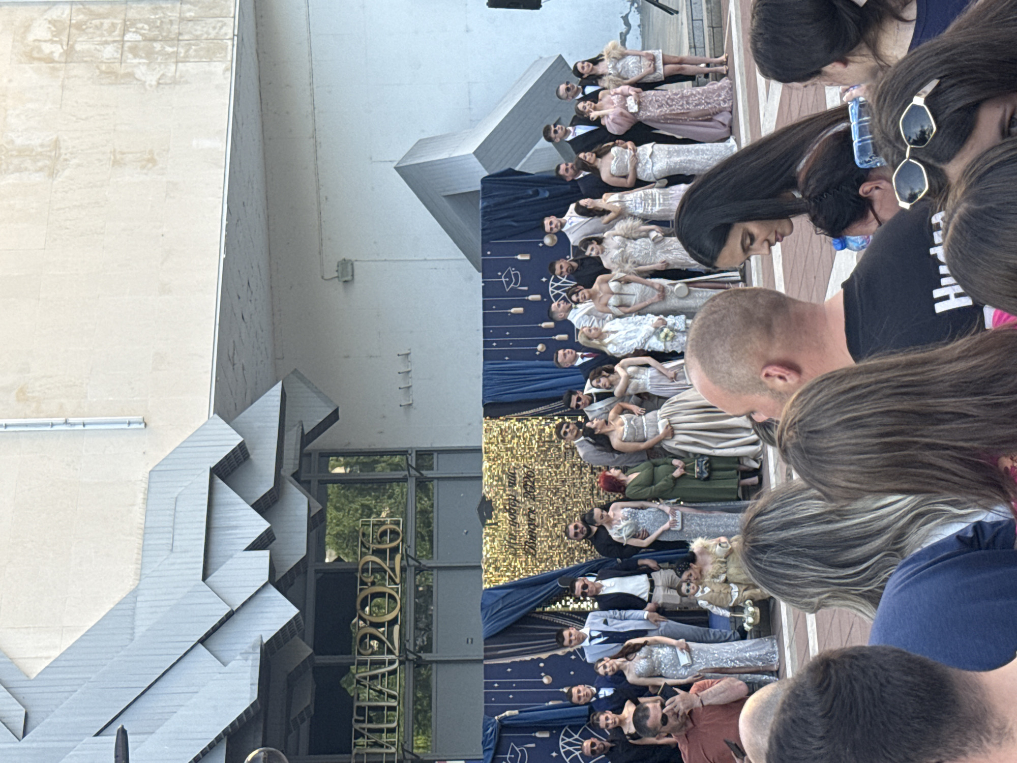 The image shows a gathering of people in Blagoevgrad, Bulgaria. In the background, a group of formally dressed individuals (both men and women) are standing on an elevated stage. The stage has a backdrop of blue and gold fabric. A sign that reads "2026" is visible on a building next to the stage. In the foreground, a crowd of people, seen from various angles, appear to be observing the group on stage.