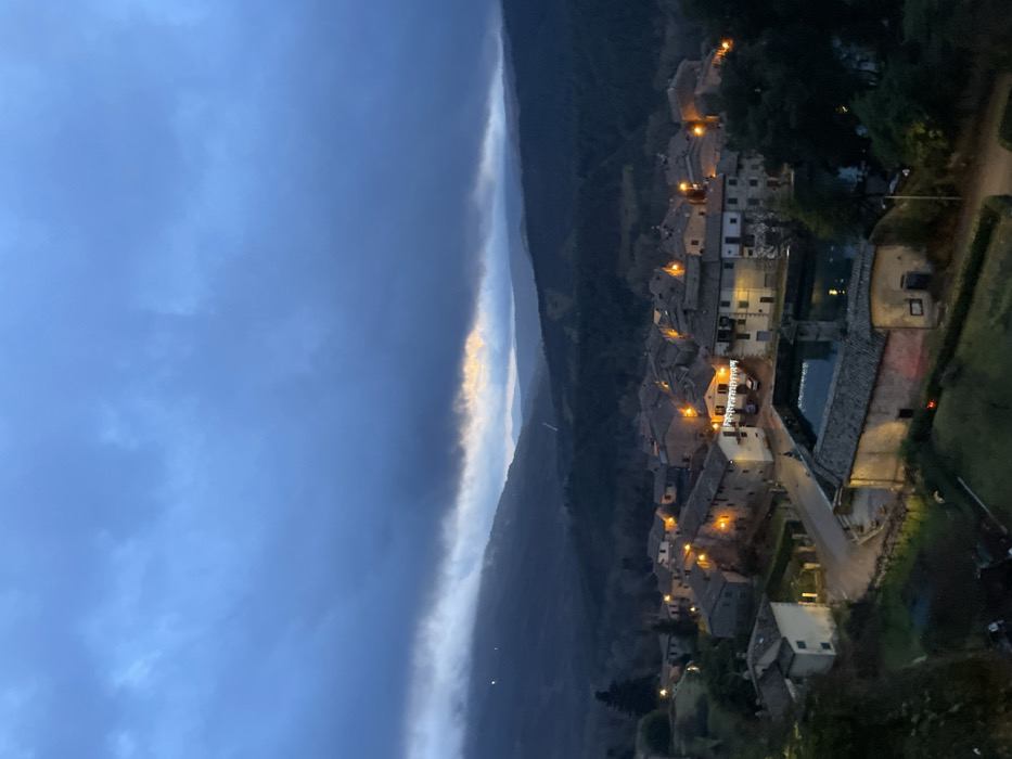 This image presents an elevated, nocturnal view of the historic town of Santa Fiora, Italy, nestled amidst undulating terrain. The scene is captured at dusk or night, indicated by the dark, overcast sky and the warm artificial illumination emanating from the buildings.
The town itself occupies the mid-right portion of the frame, showcasing a cluster of densely packed, traditional stone or stucco buildings with terracotta rooftops. Numerous streetlights and lights from within the buildings cast a warm, golden glow, highlighting the architectural details and narrow pathways. A notable detail is the illuminated sign "Ferrante" visible on one of the central buildings within the town. To the left of the town, what appears to be a body of water or a large communal basin reflects some of the surrounding light.
Beyond the town, rolling hills and mountains recede into the distance, appearing dark and silhouetted against the horizon. The sky, which dominates the upper left, is a deep, dark blue-grey, heavy with clouds. A striking feature is a distinct, narrow band of brighter, almost glowing clouds stretched across the horizon line, separating the dark sky from the distant mountains. This band, possibly catching the last remnants of twilight or a break in the cloud cover, adds a dramatic contrast to the otherwise somber sky.
No specific human activity is discernible, and the overall atmosphere is one of peaceful serenity. The image offers a tranquil glimpse of an Italian hill town as night falls.