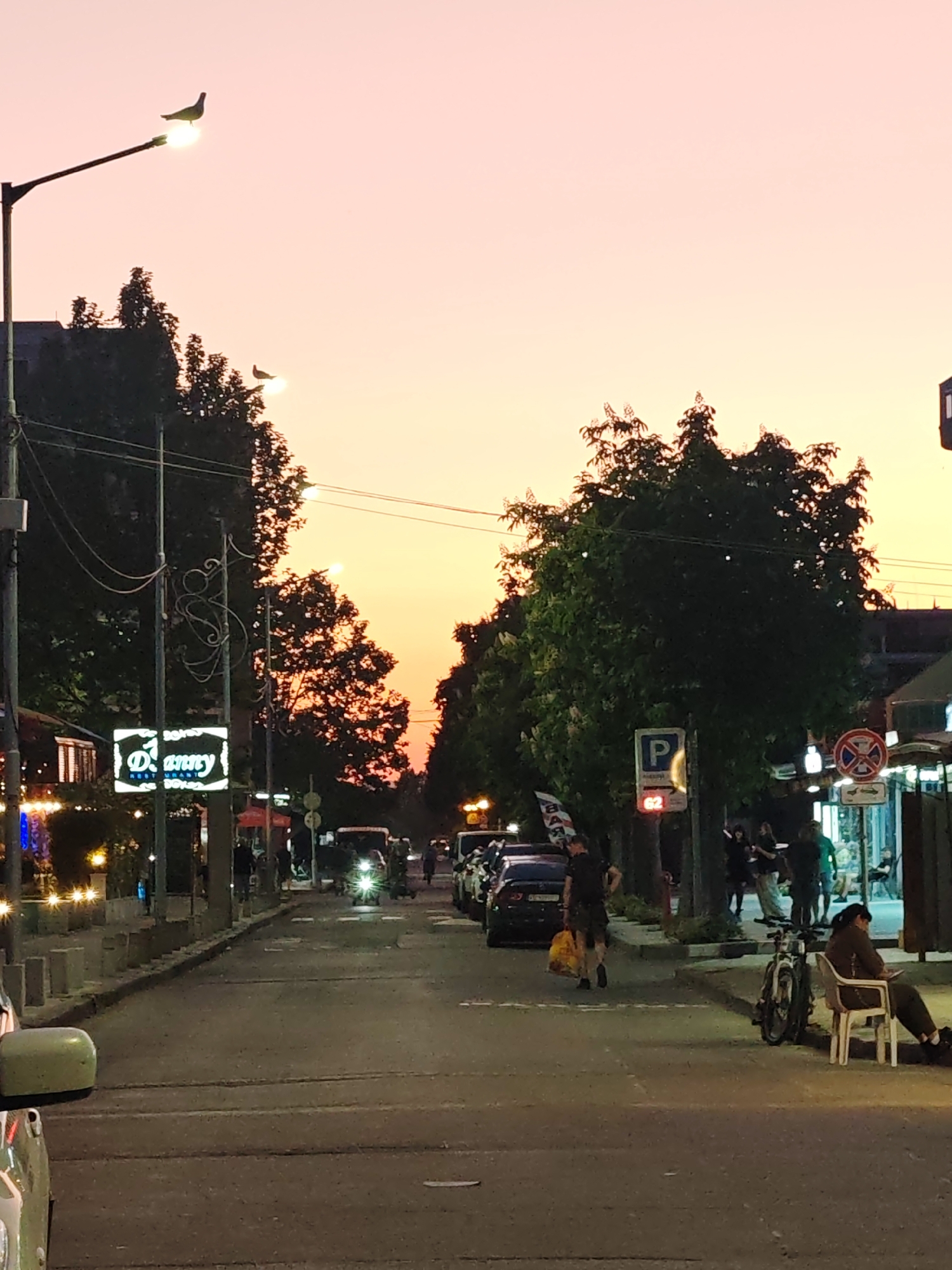 The image depicts a street scene in Nesebar, Bulgaria at dusk. The sky is a gradient of pink and orange.
People are present in the scene, including a person sitting on a white chair next to a bicycle on the right side of the street and people standing in groups or walking on the sidewalks. A man is walking down the street, carrying a yellow bag.
Several cars are parked along the right side of the street, and other cars are driving down the road.
Signs are visible, including a parking sign, a no entry sign, and a restaurant sign that reads "Danny". Streetlights illuminate the street. Trees line both sides of the road.