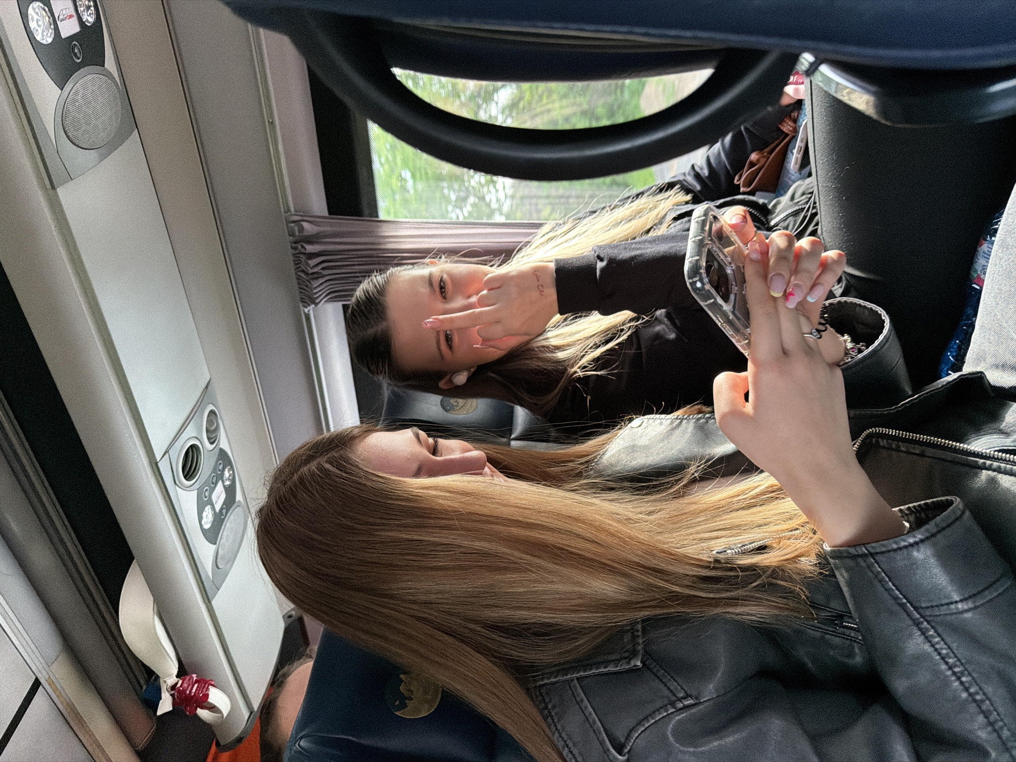 The image captures two young women inside what appears to be a bus or train cabin, likely during daytime, as indicated by the natural light filtering through the window. The scene suggests travel, possibly in Gelemenovo, Bulgaria.
In the foreground, a young woman with long, flowing light brown hair, wearing a dark leather jacket, is visible in profile. She is looking downwards, possibly resting or focused on something out of frame. Her hands, holding a cell phone in a clear case, show a detailed French manicure with a small pink heart on one nail and a blue tip on another. A silver ring is visible on her finger. A dark bottle and part of a dark seat with a golden, circular emblem are also seen on the right.
In the background, another young woman with long dark blonde hair is looking directly at the camera, making a hand gesture with her middle finger extended. She is wearing a black long-sleeved top and has a white earbud in her ear. A small tattoo, reading "دهب" (Dahab, meaning "gold" in Arabic), is visible on her inner right forearm. Her nails are also manicured with a red and white pattern. Behind her, a small brown purse is visible.
The interior of the vehicle features an overhead control panel with various buttons, lights, and a speaker. Below the window, a set of pleated blinds is partially visible, and outside, lush green foliage suggests a natural landscape. A dark, U-shaped structure, resembling a part of a seat or luggage rack, is in the upper right. A small, decorative item, possibly a ribbon in red and white, is tied to a fixture near the overhead panel on the left. The overall impression is candid and informal.