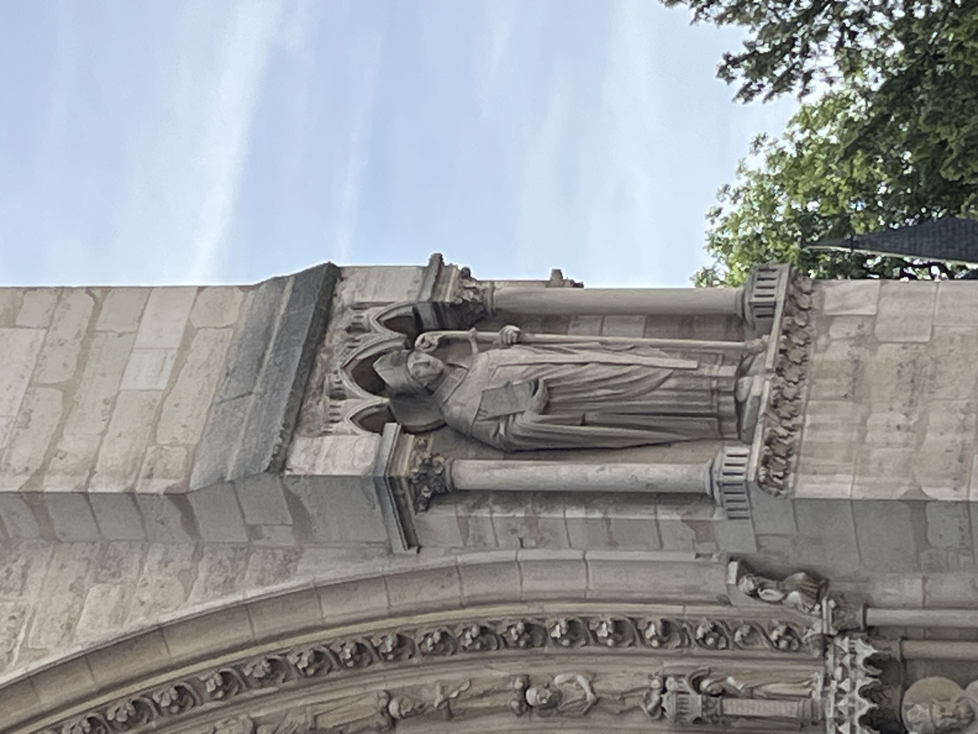This is a vertical, eye-level, medium shot of a stone statue of a robed figure holding a book and a staff, set within an architectural niche on the facade of a building. The statue is carved in a classical style, with detailed folds in the robes and a mitre on the head. The figure appears to be a religious or historical personage. The niche is supported by columns and adorned with decorative elements, suggesting it is part of a significant structure such as a cathedral or a historical monument. To the left of the niche, a portion of an arched architectural feature is visible, decorated with a repeating pattern of floral motifs. Further to the left, smaller carved figures are seen in recessed sections. The background consists of a bright, pale blue sky with faint wispy clouds, and the tops of lush green trees are visible on the right side of the frame. The architectural style suggests this is likely Notre Dame Cathedral in Paris, France.