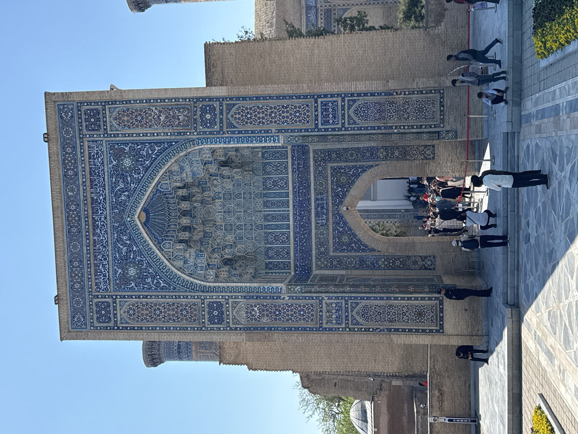 The image shows a large brick building with intricate blue and white tilework, located in Samarkand City, Uzbekistan. The building is likely a mosque or mausoleum, as evidenced by the architectural style and design. A group of people are standing near the entrance of the building, some waiting in line. The ground in front of the building is paved with large stone tiles. The sky is clear and blue.