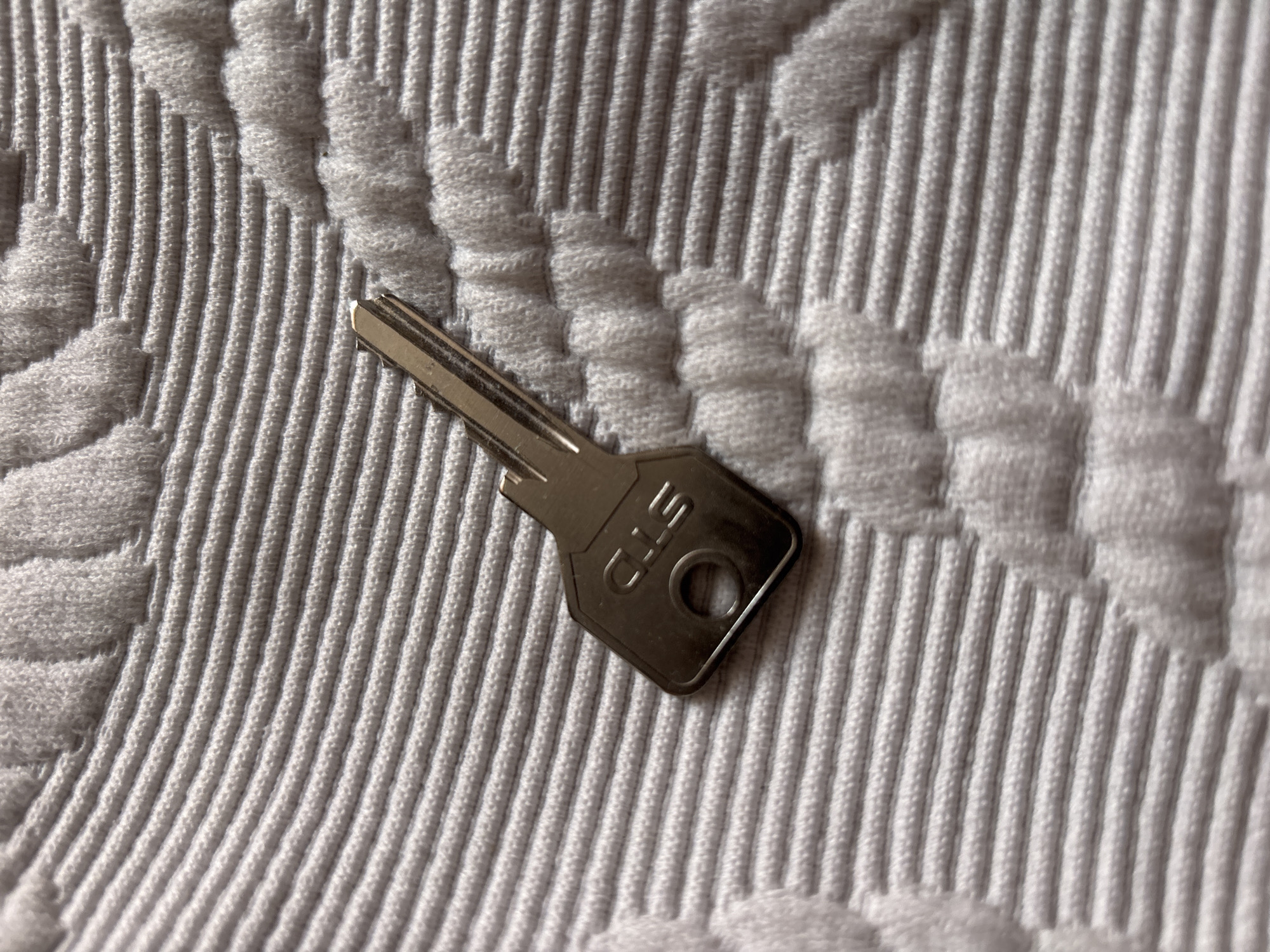 A single, silver-colored metal key with the etched letters "STD" is lying diagonally on a textured, white fabric surface. The fabric has raised, ridged patterns. The key is positioned with its head on the lower right and its shaft extending towards the upper left. The lighting casts subtle shadows, highlighting the texture of the fabric and the key. There are no people, animals, or other objects present. The image appears to be a close-up shot of the key on the fabric.
