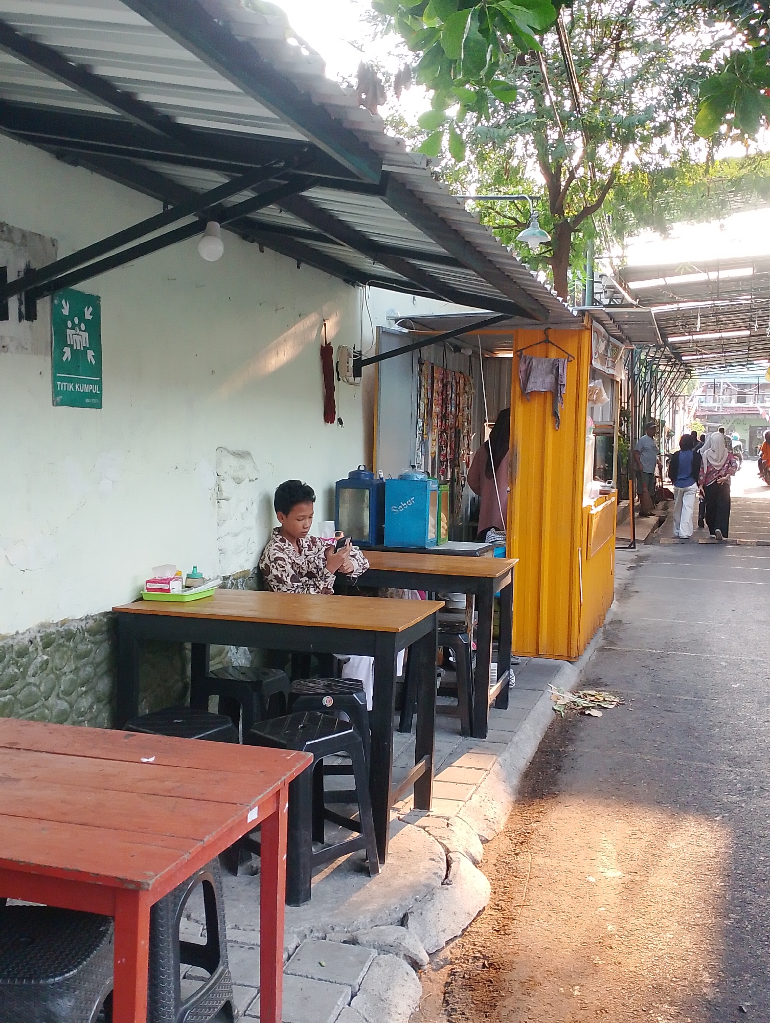 A young boy, dressed in a patterned shirt, sits alone at a table in an outdoor food stall area. He is looking at his mobile phone. In front of him on the table is a green tray with tissues and small containers. Two black stools are placed under the table. To the left of the boy's table, another red table is visible. Behind the boy, a blue water dispenser is present. The stall is constructed with a yellow corrugated metal structure and a metal roof. A sign with green figures and the text "TITIK KUMPUL" (meeting point) is mounted on the wall. In the background, other stalls line a pathway, and several people are walking by. The scene appears to be in a street market or hawker center in Yogyakarta, Indonesia, given the presence of the "Titik Kumpul" sign, which is often used for emergency assembly points in Indonesian public spaces. The lighting suggests it is late afternoon.
