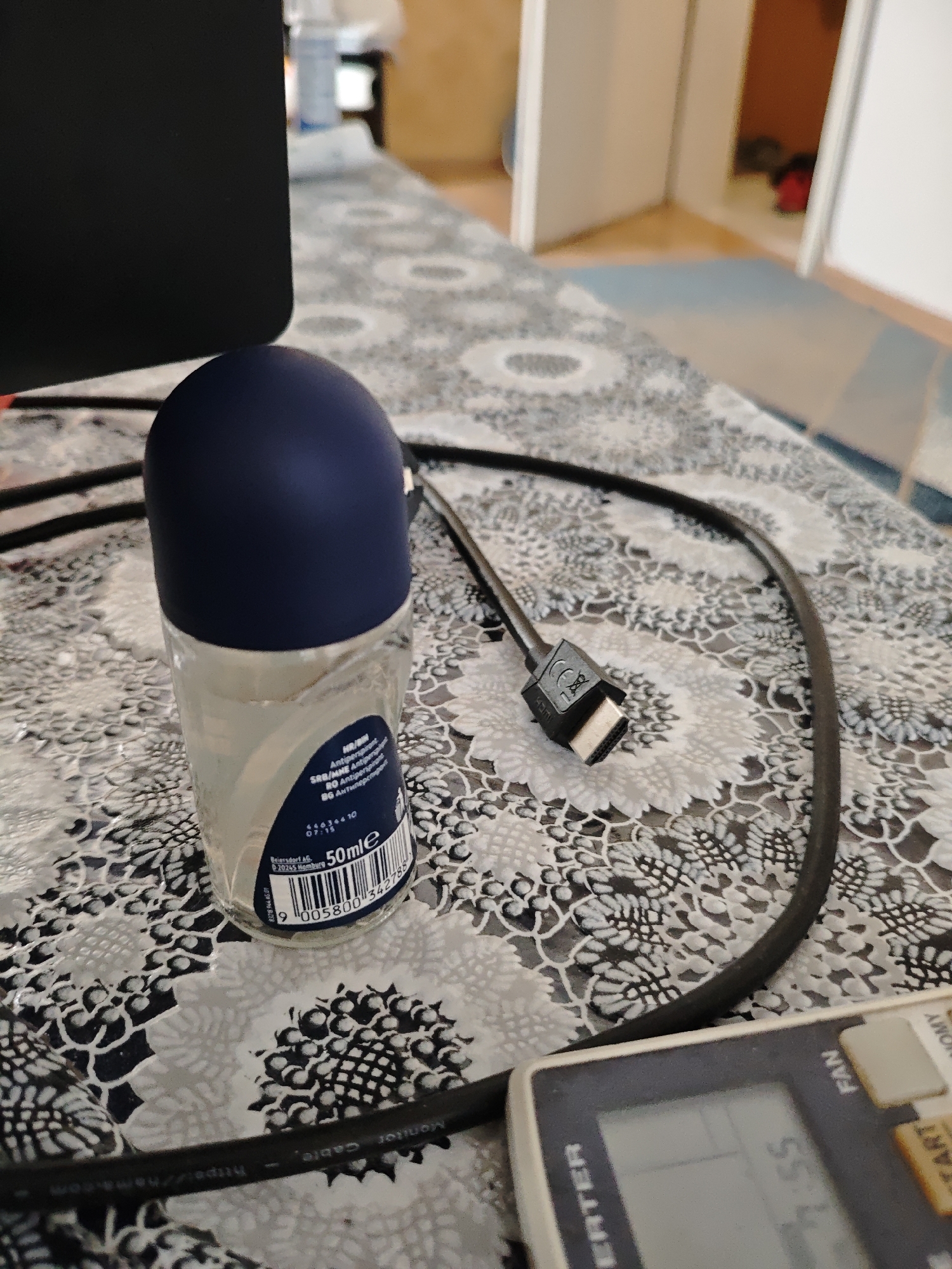 The image depicts a close-up, top-down view of a roll-on deodorant bottle with a dark blue cap, resting on a patterned tablecloth. A black HDMI cable is also visible, with its connector end resting near the deodorant. In the lower right corner, a portion of a digital device, possibly a remote control, is visible, displaying some text and numbers. The background is out of focus but suggests an indoor environment with furniture and doorways. The tablecloth has a white floral or lace-like pattern on a dark background. The scene is likely set on a table or desk in a domestic or office setting. There are no people visible in the image. The location in the city of Plovdiv, Bulgaria, is not determinable from the image content.