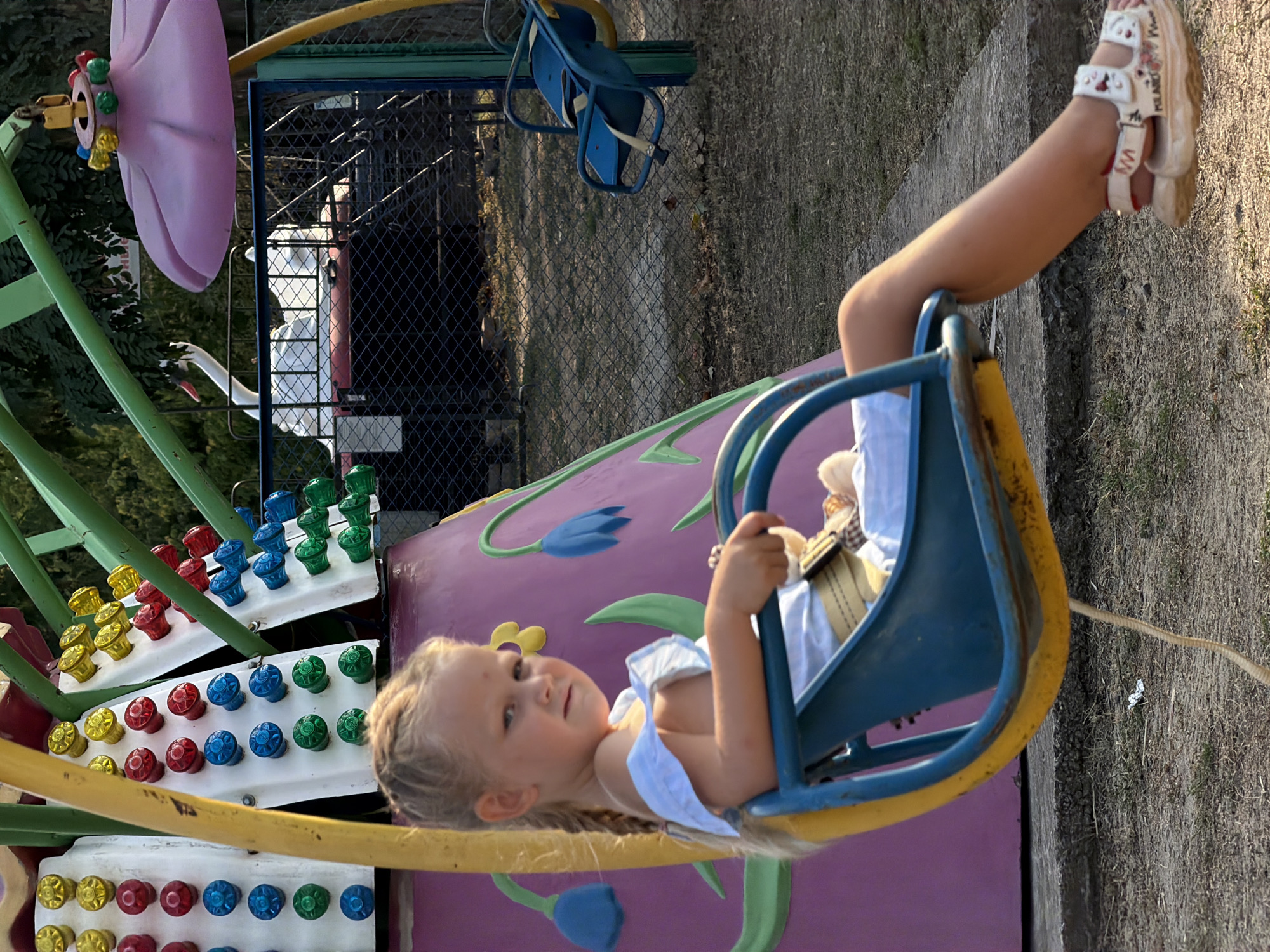 A young Caucasian girl with blonde braided hair sits in a blue and yellow swing. She is wearing a light blue off-the-shoulder dress and white sandals. She is holding a small stuffed toy. The swing is part of a playground ride with a purple base adorned with green and blue floral decorations. In the background, a white swan-shaped ride is visible behind a black metal fence. The scene is outdoors, with trees and dry grass. The location is identified as Kamyshin, Russia.