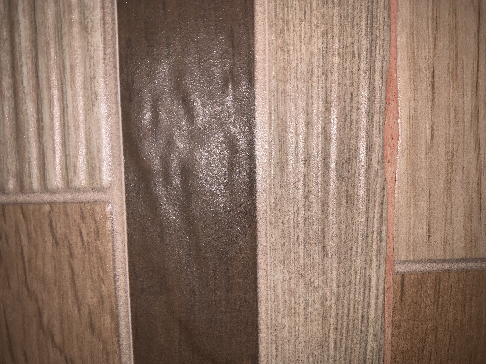 The image presents a close-up, abstract view of an interior surface, likely a wall or floor, composed of decorative tiles or panels. The setting is a part of a structure, possibly a home or commercial building, given the location context of Santa Ana, Venezuela.
The surface is characterized by a pattern of vertical strips in varying shades of brown and beige. A prominent central vertical strip is dark brown and features a glossy, somewhat undulating texture that reflects light, creating bright, scattered highlights across its surface. Flanking this central strip are panels exhibiting a lighter, beige-brown wood-grain pattern with distinct vertical striations, suggesting a natural wood aesthetic. These wood-grain sections appear to have a more matte or slightly textured finish compared to the glossy central strip.
The individual panels and sections are separated by thin grout lines. Some of these lines, particularly the vertical ones between the wood-grain patterned panels, are a distinct reddish-pink or terracotta color. Horizontal grout lines also divide the vertical panels into larger rectangular sections, creating a tiled or paneled effect. The lighting appears to be artificial, accentuating the different textures and reflective qualities of the materials. There are no people, activities, events, or visible text in the image. The overall impression is one of contrasting textures and earth-toned colors, contributing to a decorative architectural element.