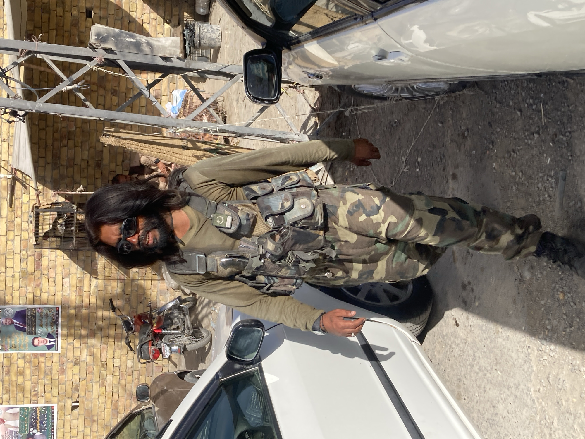 The image shows a man wearing camouflage clothing and sunglasses standing between two white cars in an urban environment. He has long dark hair and a beard. He is wearing a vest with multiple pouches. A motorcycle is visible parked against a brick wall, which has posters on it. Overhead, there is a metal structure and a brick building. The ground appears to be concrete or asphalt. Based on the information, the image is likely from the city of Quetta City Tehsil, Pakistan.