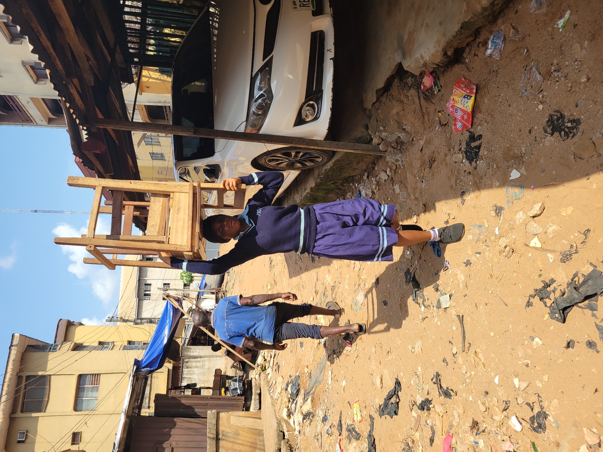 A young person wearing a navy blue sweater and blue shorts with light blue trim is carrying a wooden chair on their head. The person is walking on a dirt path strewn with litter, including black plastic bags, wrappers, and other debris. In the background, there is a white car parked under a corrugated metal overhang. Behind the car, a multi-story building can be seen. To the left of the frame, another person in a blue t-shirt and dark pants walks away from the camera. The scene appears to be in a somewhat dilapidated urban area in Onitsha, Nigeria, characterized by the presence of buildings, vehicles, and the general state of the ground. The lighting suggests it is daytime.