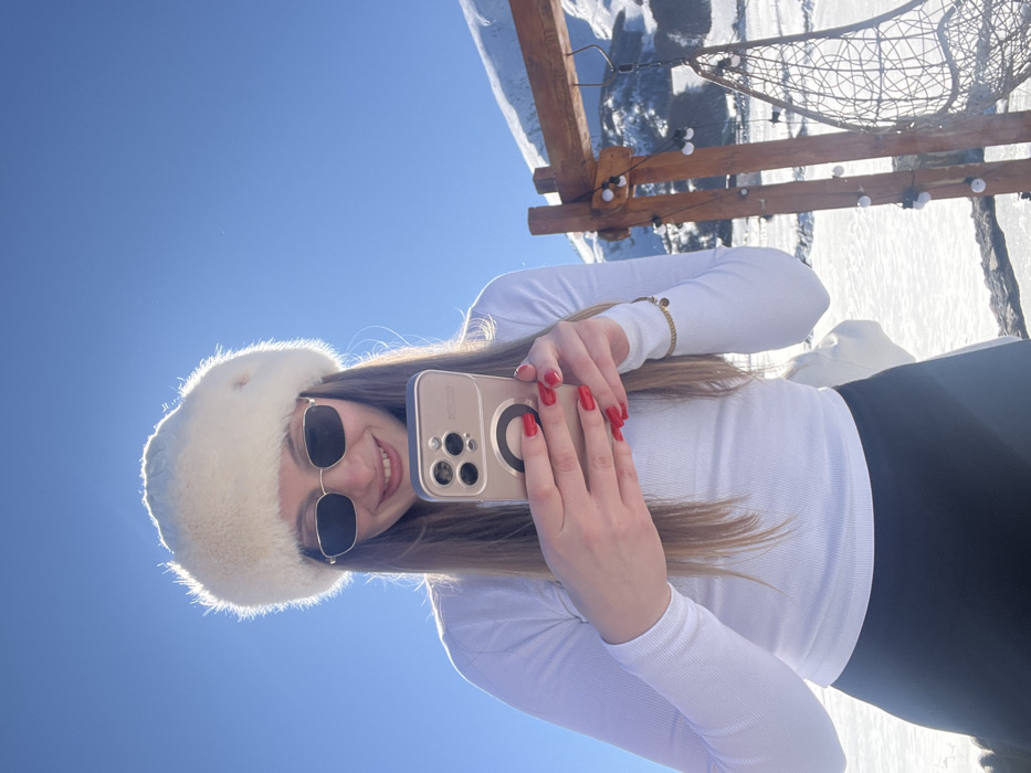 The image depicts an outdoor winter scene in Virak, Montenegro, under a clear blue sky. A young adult female is centrally positioned, smiling and looking directly towards the viewer. She wears a white, fluffy winter hat, dark rectangular sunglasses, a white ribbed long-sleeved shirt, and dark trousers. Her long, light brown hair is visible. She holds a light-colored smartphone, featuring a black circular attachment on its back, in both hands at waist height. Her fingernails are painted bright red, and a gold-colored bracelet with a small charm is visible on her right wrist. In the background, a snow-covered landscape extends, culminating in distant snow-capped mountains. To the subject's right, a rustic wooden frame structure, wrapped with string lights composed of white bulbs, is visible. A wicker-style hanging chair is suspended from this structure. The ground is uniformly covered in snow.