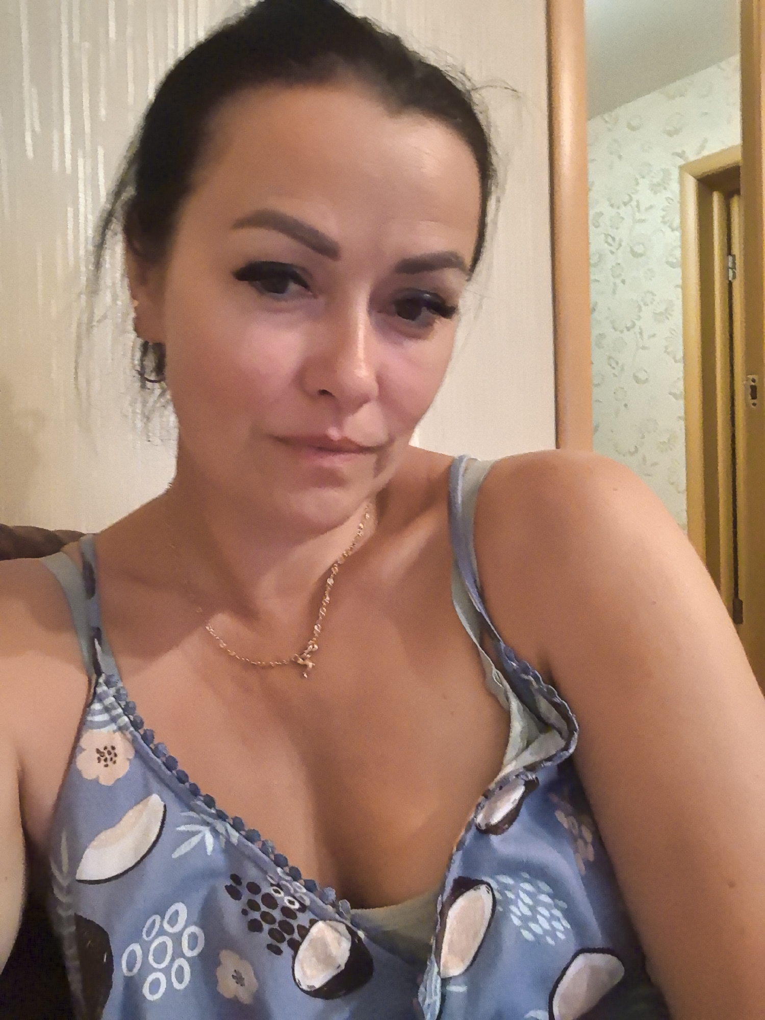 The image shows a close-up, selfie-style shot of a woman. She has dark hair pulled back, prominent eyebrows, and is wearing makeup, including eyeliner and false eyelashes. She is also wearing a blue floral-patterned dress with a beaded trim. A delicate gold necklace with a small charm is visible around her neck.
The background consists of textured wallpaper and a wooden door frame, suggesting an indoor domestic setting. There is a hint of a doorway or another room visible in the background, with a door that appears to be closed. The lighting is warm and seems to come from artificial sources.
It is not possible to determine the location in the city of Lida, Belarus from the visual information present in the image.