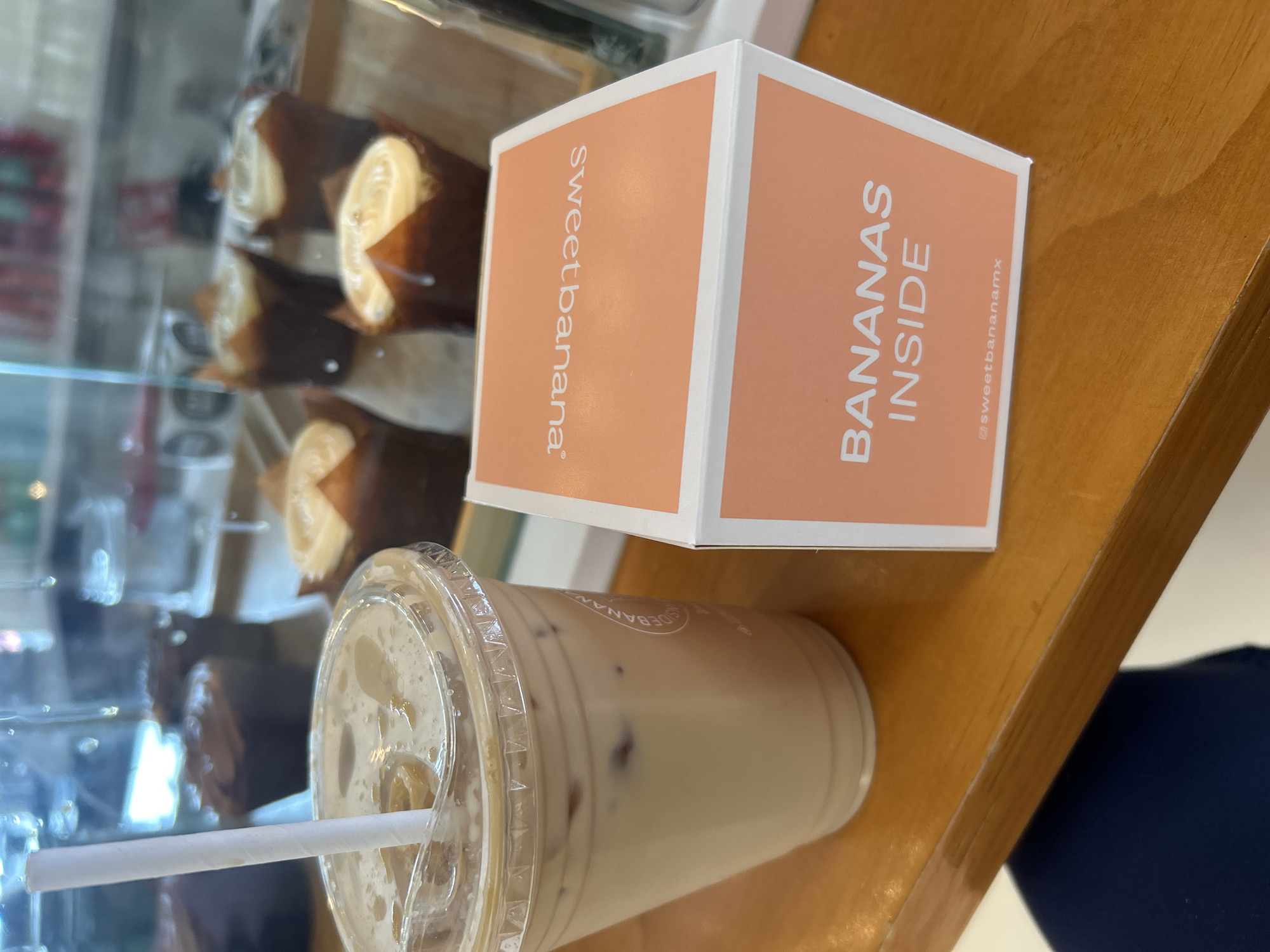 The image displays an indoor scene, likely a cafe or dessert shop, featuring a packaged product and a beverage on a light brown wooden counter or table.
The main subjects are a small, rectangular box and a plastic cup with a straw. The box is light orange with white edges. On its visible front panel, the text "BANANAS INSIDE" is printed vertically in large, dark brown capital letters, with "@sweetbananamx" in smaller text below it. The adjacent side panel of the box reads "sweetbanana®" vertically in dark brown lowercase letters.
Next to the box is a clear plastic cup with a lid and a white straw. The cup holds a light, milky brown liquid, suggesting an iced coffee or similar beverage, with small, darker specks possibly being ice or syrup. Faint branding is visible on the cup, partially obscured, appearing to be related to the "sweetbanana" brand.
In the blurred background, behind a reflective glass display case, several individual baked goods or desserts are visible. These items are presented in dark brown paper liners or wraps, showcasing a light, creamy-colored interior or topping. They appear to be small, possibly banana bread portions, rolls, or mini cakes, consistent with a "sweetbanana" theme.
No people are visible, and no specific activity is taking place other than the presentation of these food and drink items. The lighting is consistent with an indoor setting during daytime operating hours.