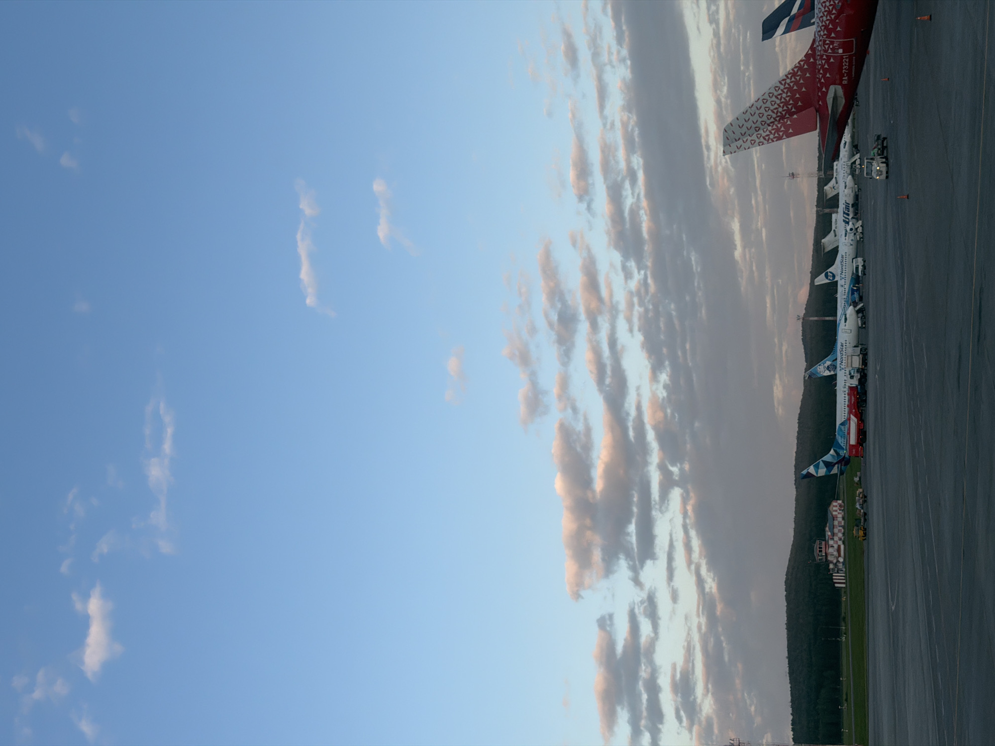 The image depicts an airfield at dusk in Emelyanovo (airport), Russia. Several airplanes are parked on the tarmac. In the foreground, the tail of a red airplane with a distinctive white triangular pattern and registration "RA-73221" is prominent. Further down the tarmac, other aircraft are visible, including one with "NordStar" branding and another with "Utair" livery. In the background, a control tower with red and white markings stands near a wooded hill. The sky is a blend of blue and scattered clouds tinged with the warm colors of sunset. The overall scene suggests a quiet moment at the airport before nightfall, with ground crew vehicles and orange traffic cones present on the tarmac.