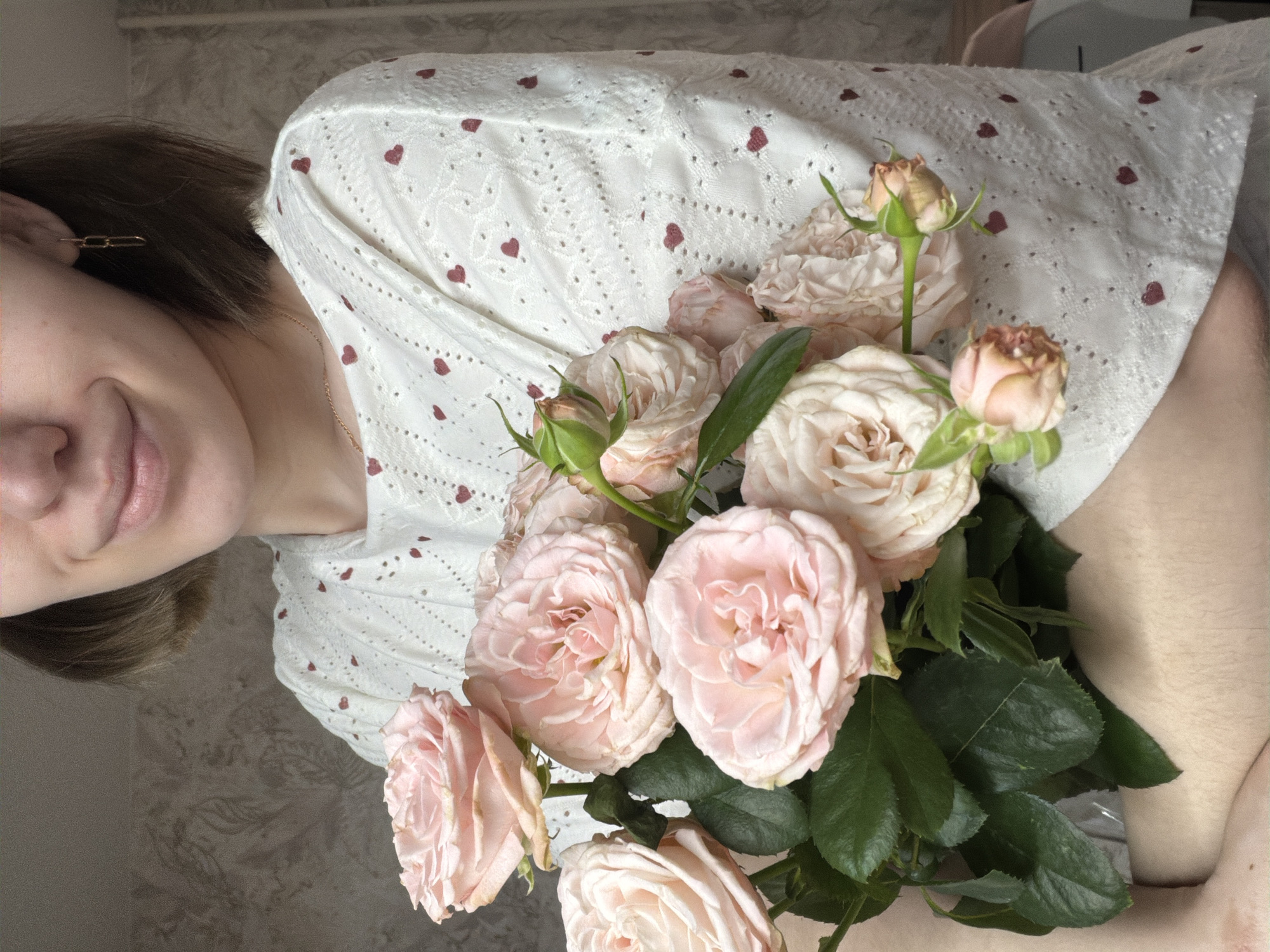A woman holds a bouquet of pale pink roses with green foliage. She is wearing a white blouse with small red hearts. She is smiling slightly and wearing a gold chain necklace and a gold earring. The background appears to be a wall with a subtle patterned wallpaper. The location is not discernible from the image.