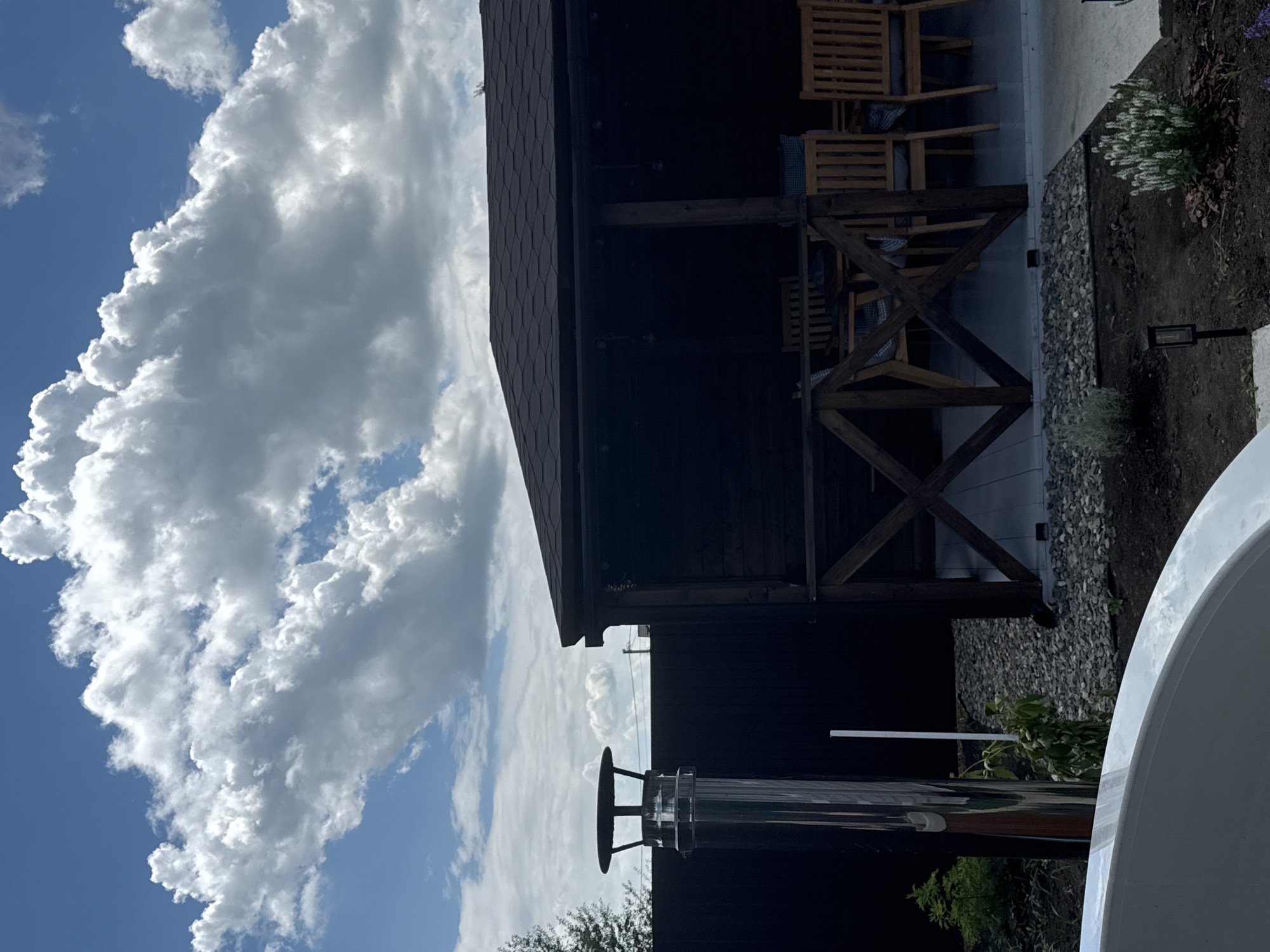 The image depicts a backyard scene on a sunny day with scattered clouds. In the foreground, the curved edge of a white bathtub or hot tub is visible. To the left, a tall, shiny metal chimney pipe stands vertically. Behind the chimney is a dark wooden fence. Further back, a dark wooden gazebo with a brown shingled roof is present. The gazebo has an open front with a wooden railing. Inside the gazebo, wooden chairs and tables are arranged. The ground around the gazebo is covered with gray gravel and dirt, with some small plants and gardening lights. The sky is blue with large, fluffy white clouds. The scene appears to be located in a suburban or rural residential area in Starokorsunskaya, Russia, given the overall aesthetic of the landscaping and outdoor structures. There are no people visible in the image.