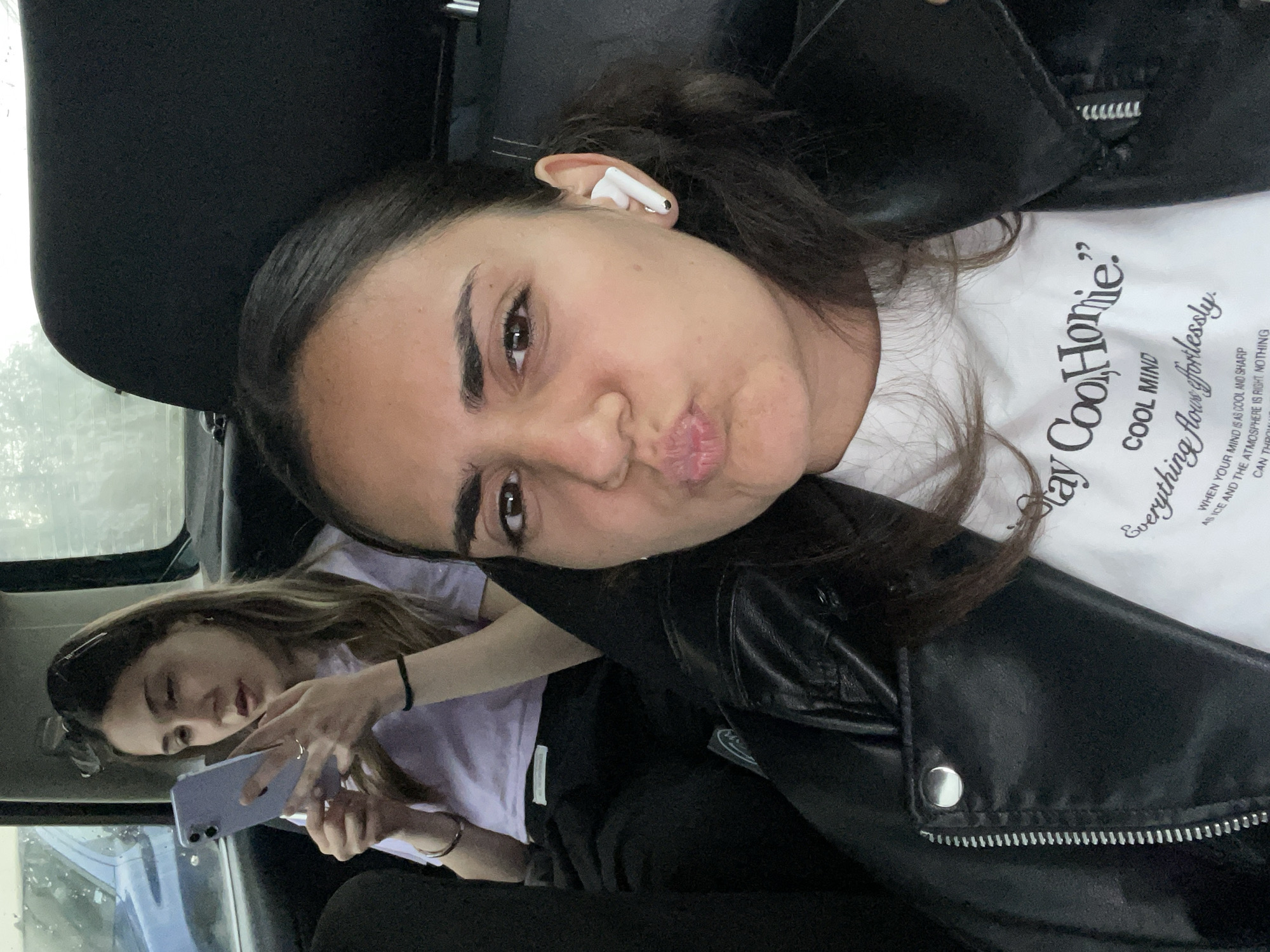 Two young women are in the back of a vehicle. The woman in the foreground is taking a selfie. She is wearing a white t-shirt with writing and a black leather jacket. She is wearing an earbud in her ear and making a kissing face. The woman in the background is looking at a cell phone. She is wearing a light purple t-shirt. The interior of the vehicle is dark with black seats and a black dashboard. The location is not confirmed to be in Debelt, Bulgaria, as there are no identifiable contextual clues.