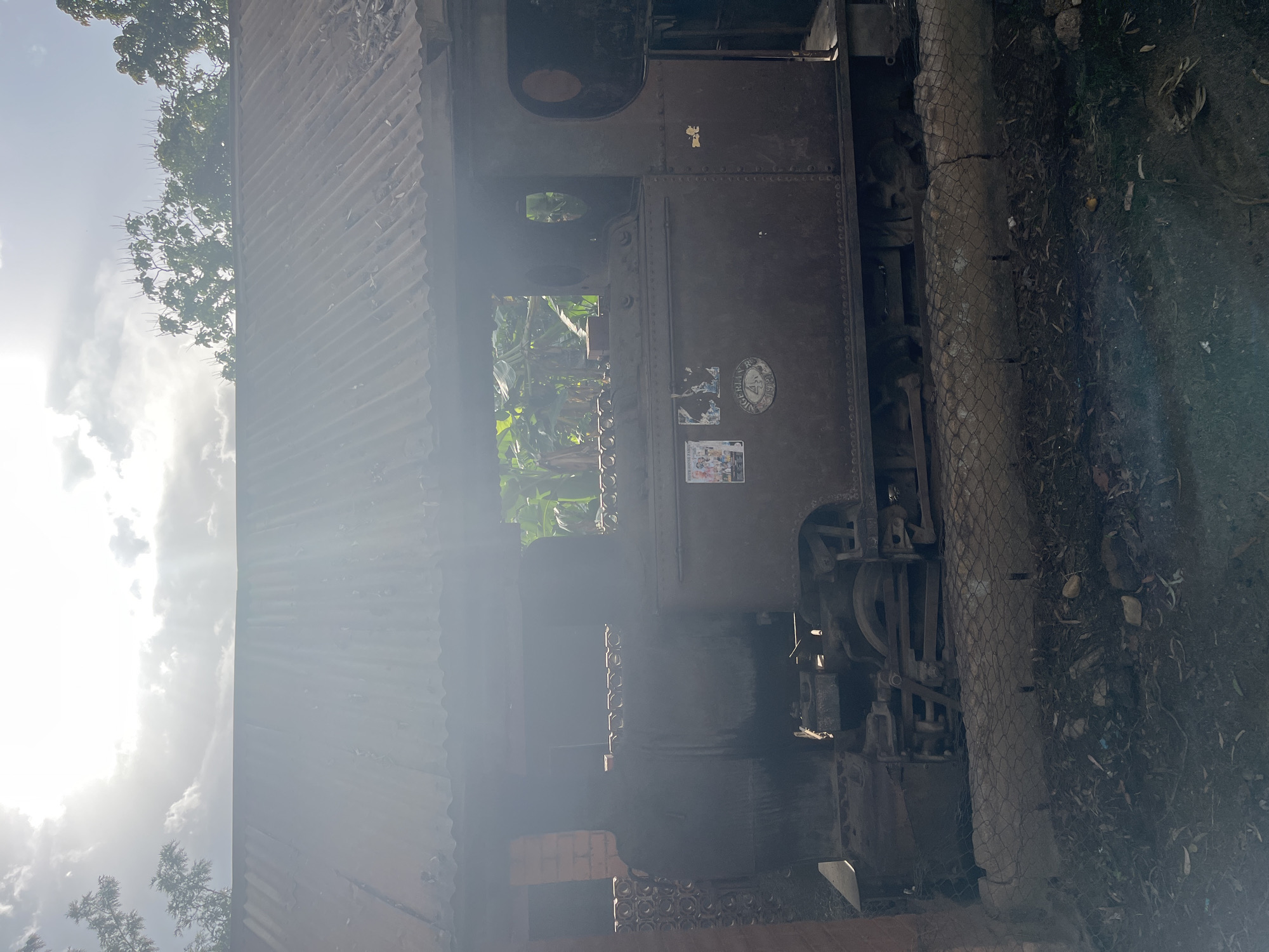 An abandoned, rusty steam locomotive is housed under a corrugated metal roof in Jos, Nigeria. The train appears to be from the colonial era, with "NIGERIAN RLY 492" emblazoned on its side. The locomotive is partially covered by a brick structure on the left, and vegetation is visible through the open windows of the train's cabin. The surrounding area is unkempt, with debris and dry leaves scattered on the ground, suggesting the site has been neglected. The sky is bright with visible sun rays, indicating it is daytime.