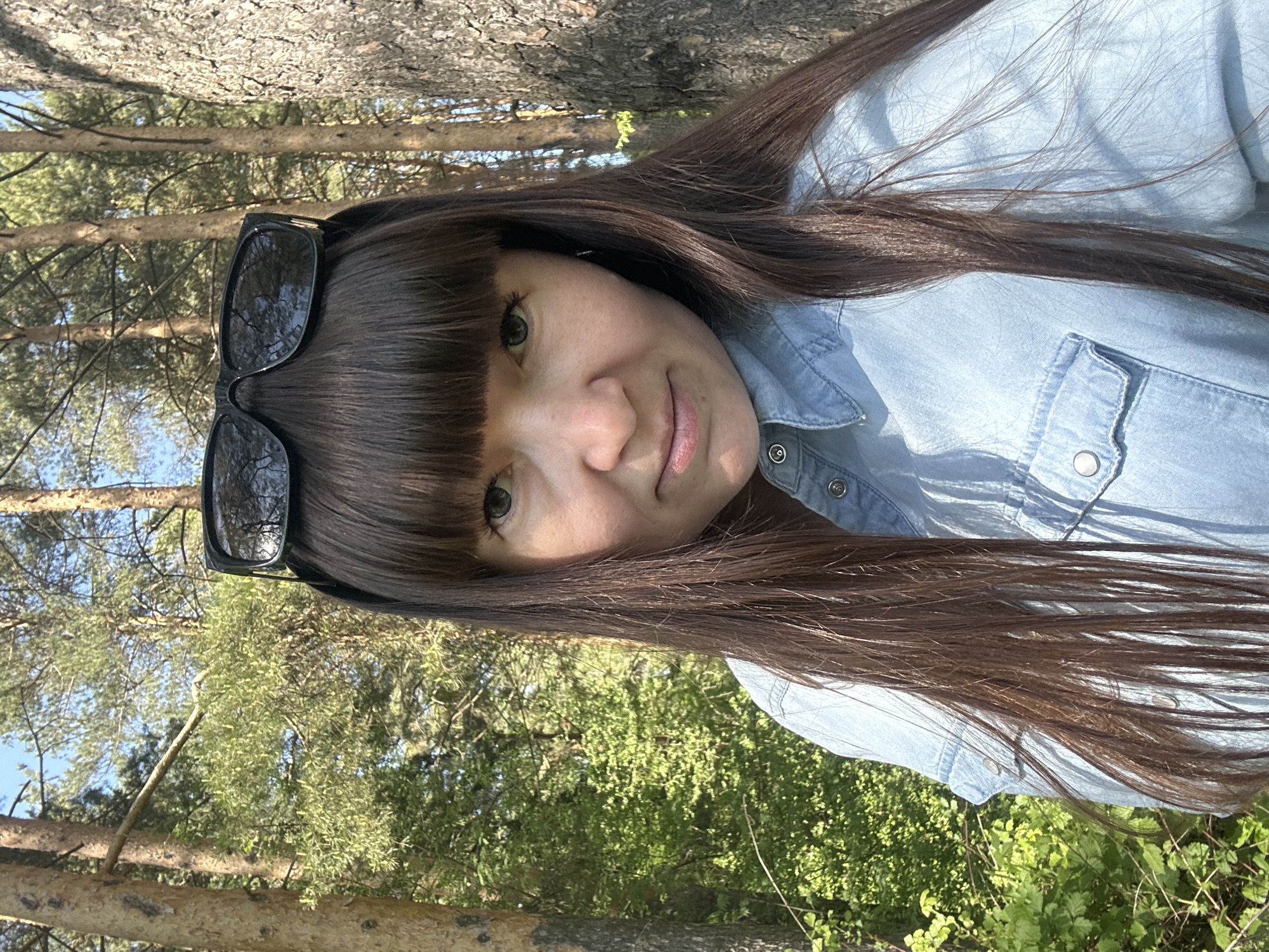 A woman with dark brown hair and bangs is pictured outdoors in daylight, possibly in a park or forested area. She is wearing a light blue denim shirt, and sunglasses are perched on her head. The background shows trees and greenery. The location is the city of Chaikovsky, Russia, based on the geographic location of the image. There are no apparent actions or interactions.