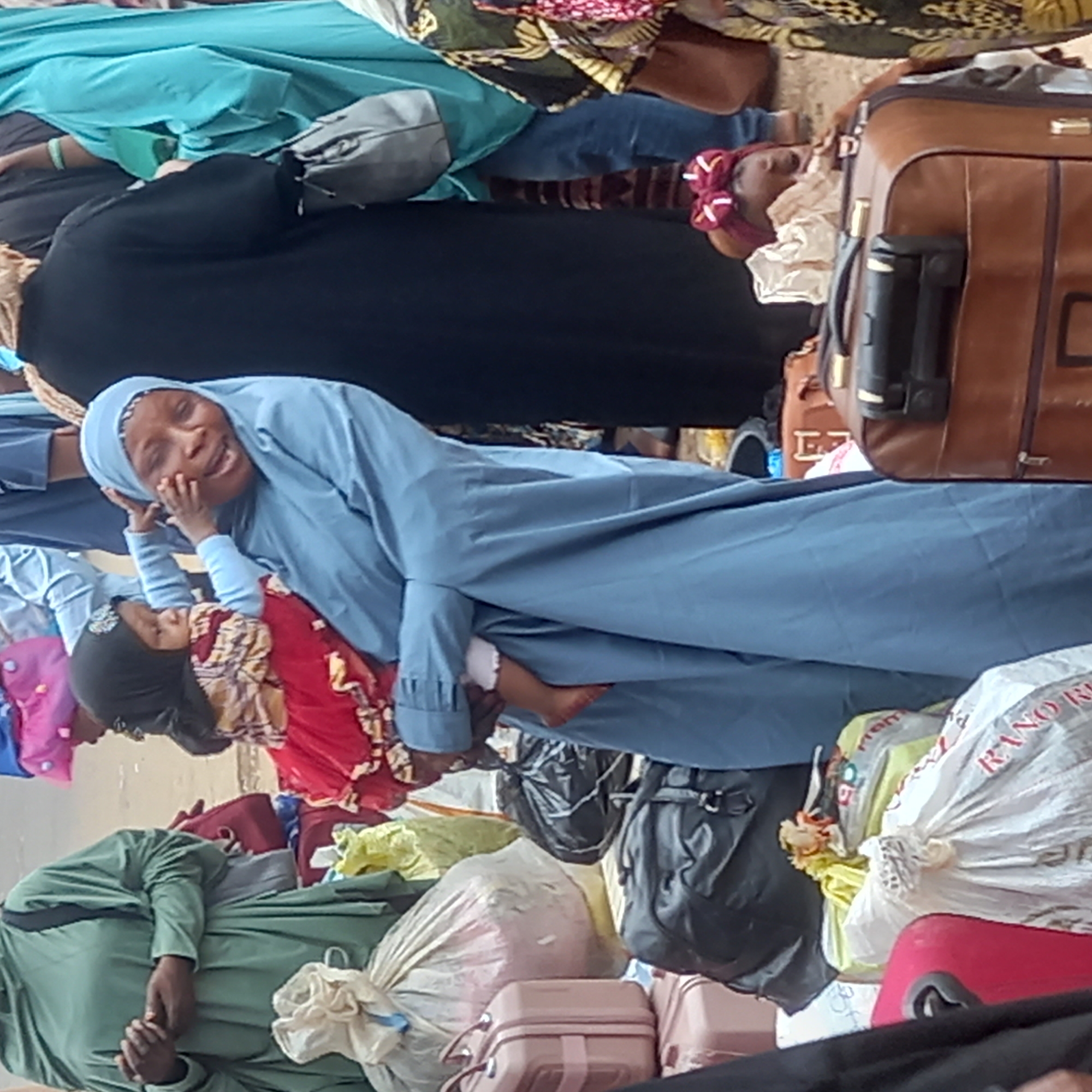 In Sardauna, Nigeria, a group of people stand amongst bags and luggage. A woman wearing a blue hijab holds a child in a red dress. She is looking at the camera and holding the child's hand to her own face. A brown suitcase stands to the right. Other people and bags are scattered throughout the scene.