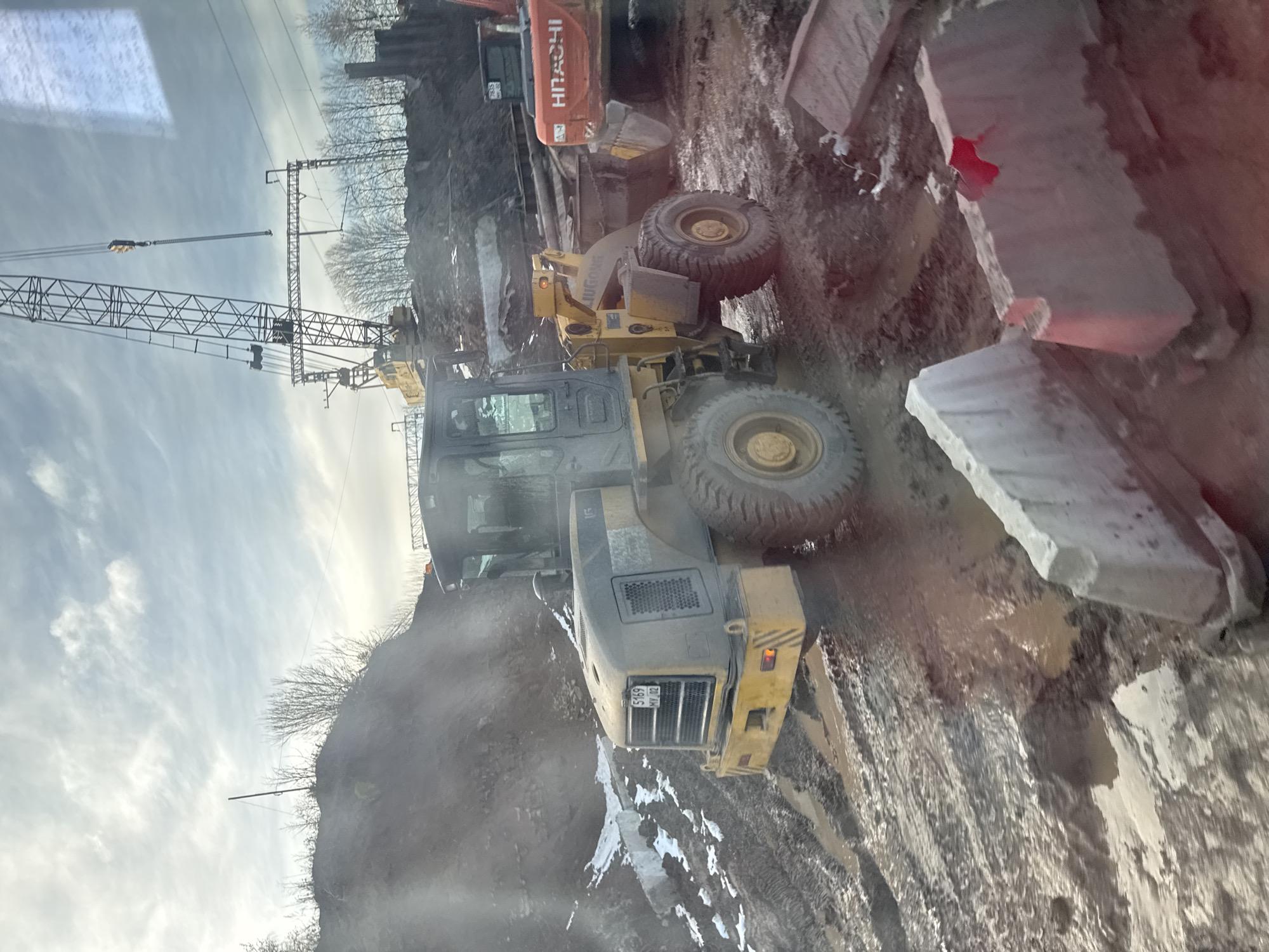 The image shows a construction site in what appears to be the city of Perm, Russia. Heavy machinery is present, including a crane, a wheel loader (possibly a LIEBHERR model), and an excavator (possibly a HITACHI). The ground is muddy and uneven, with scattered rocks and building materials. The sky is overcast. License plates from the vehicle near the front have the number 5169 MV 59 REG, with the abbreviation REG likely identifying it as a vehicle registration in Perm Krai, a Russian Federal Subject whose administrative capital is Perm.