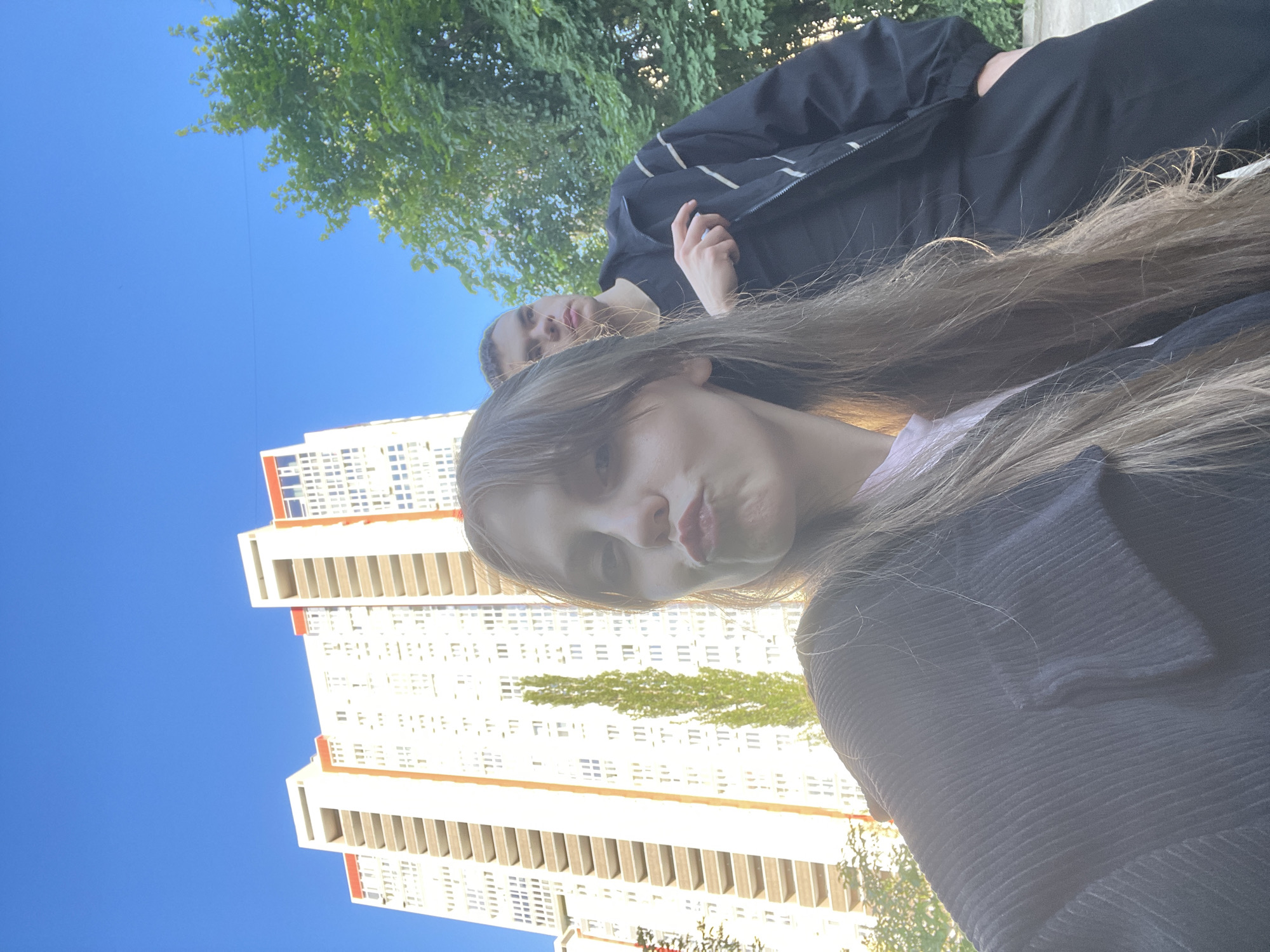 The image shows two young people in an outdoor setting, with a tall, white apartment building in the background. The sky is a clear, bright blue, suggesting it is daytime with good weather. The building has rows of windows and balconies, characteristic of Soviet-era architecture, and some orange accents around its upper floors.
In the foreground, a young woman is looking directly at the camera with her lips pursed. Her long, brown hair falls around her face and shoulders. She is wearing a dark, ribbed top. Behind her and to her left, a young man is visible. He is wearing a black jacket with white stripes and appears to be looking away from the camera. His right hand is raised, resting on the lapel of his jacket.
The image is taken from a low angle, looking up towards the subjects and the building. The overall impression is casual and candid, perhaps a selfie taken by the young woman. There are trees with green foliage visible in the upper part of the image, indicating a possibly warm season. The lighting suggests it is likely midday or afternoon, with the sun casting some bright highlights. The scene appears to be in an urban environment, given the presence of the apartment building. No text is visible in the image.