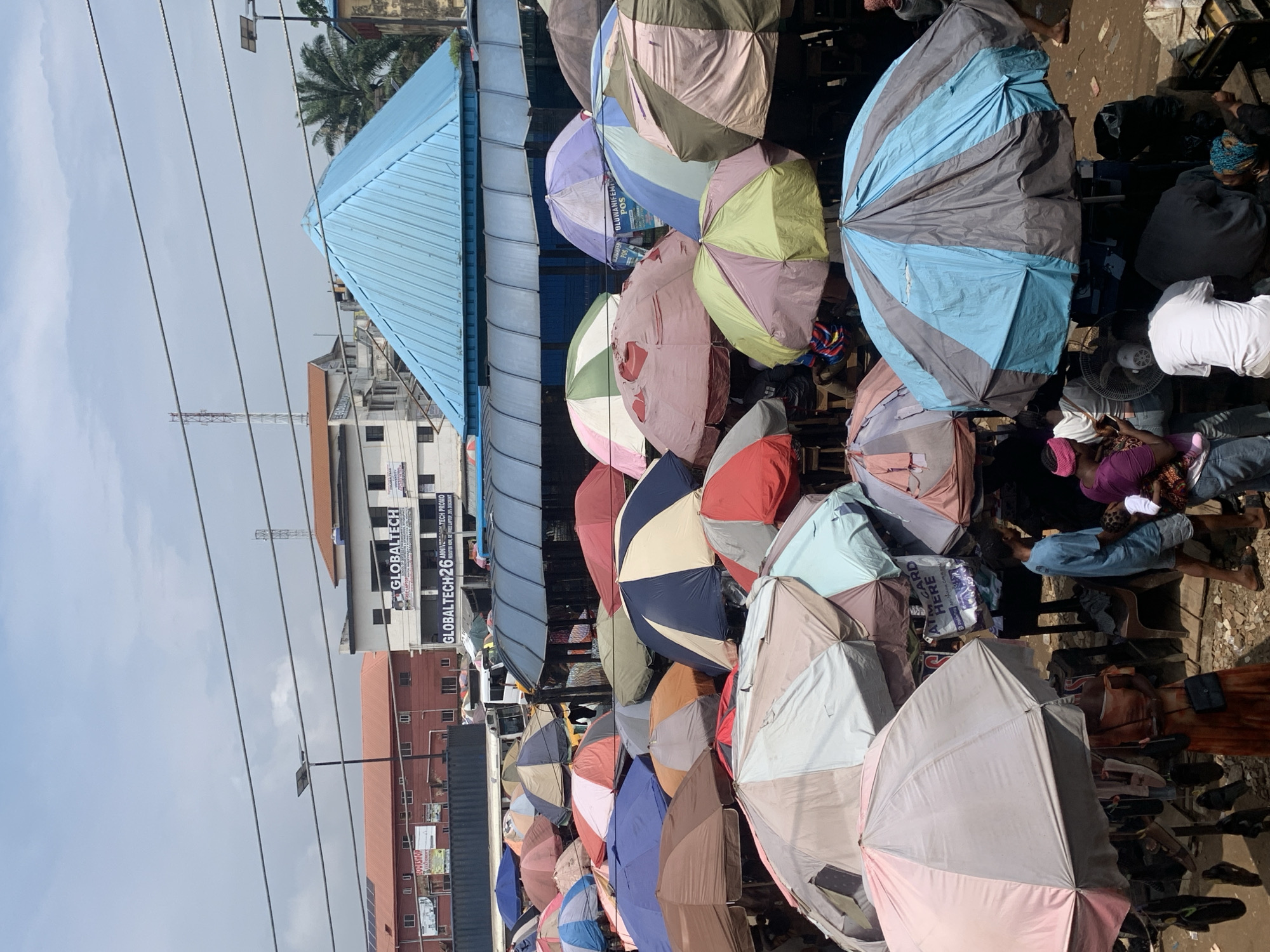 This is an overhead shot of a bustling market in Ikorodu, Nigeria. The scene is dominated by a multitude of colorful umbrellas providing shade over stalls. Numerous people are visible in the image, some actively engaged in commerce, while others are waiting or observing. The market appears to be set up on uneven, dusty ground. In the background, buildings are visible, including structures with the "GLOBALTECH 26" signage, suggesting proximity to commercial establishments. The sky is overcast with a few power lines crisscrossing overhead. The overall impression is one of vibrant, outdoor commerce within a dense urban environment.