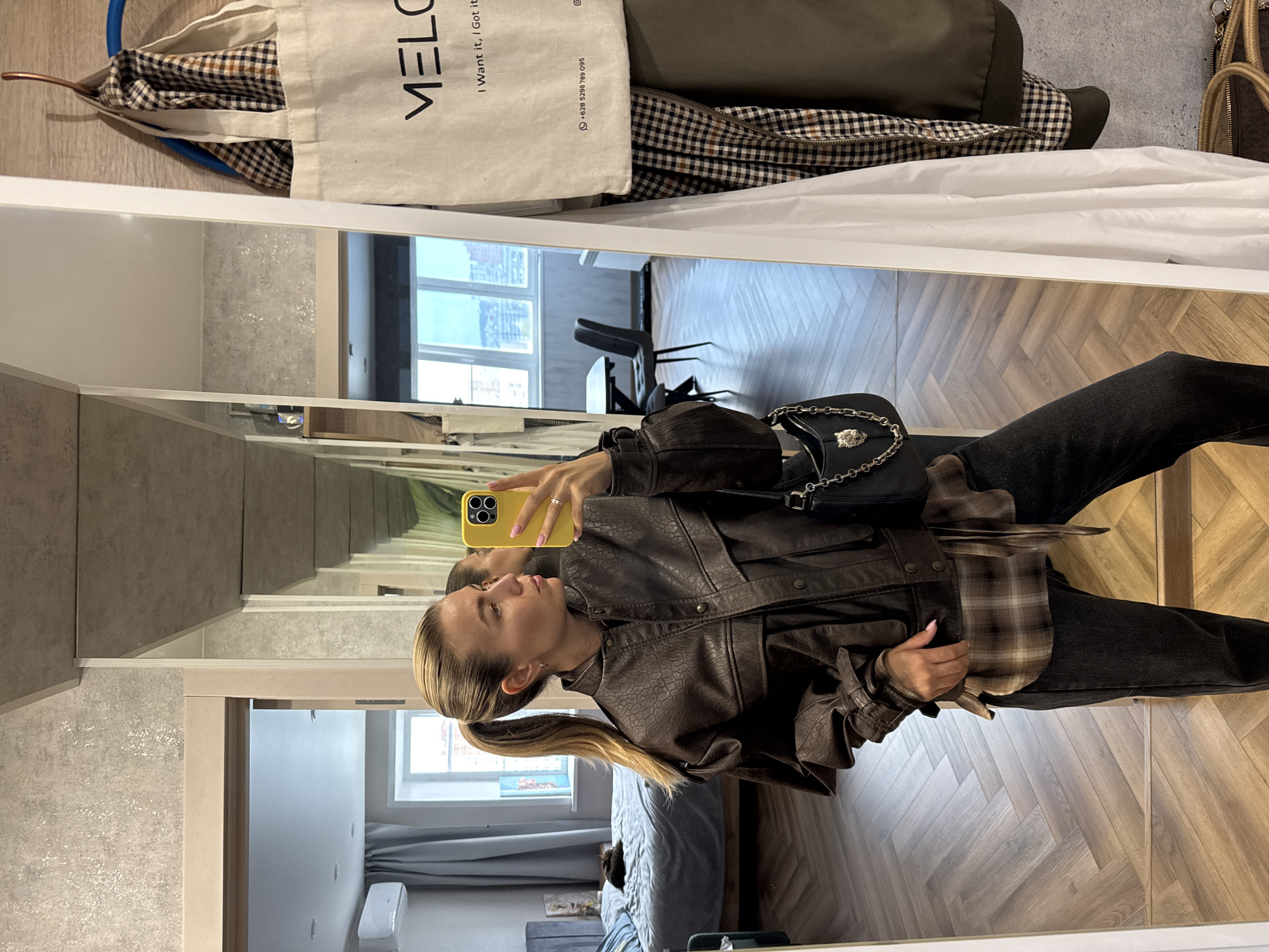 A woman with blonde hair in a ponytail takes a mirror selfie in a room that appears to be a bedroom with an adjacent dressing area. She is wearing a brown faux leather jacket over a brown plaid shirt and dark wash jeans. She is holding a yellow phone in her right hand and a black purse with a silver chain strap is on her left shoulder. In the background, a bed with blue bedding is visible, and a window with a view of a city skyline is also seen. A mirror to her right reflects more of the room, including a table and chairs. A white canvas tote bag hangs on a hook on the wall next to the mirror, displaying the word "MELO" in black lettering. A dark green bomber jacket hangs on a hanger on the wall next to the tote bag. The floor is covered with a herringbone pattern hardwood.