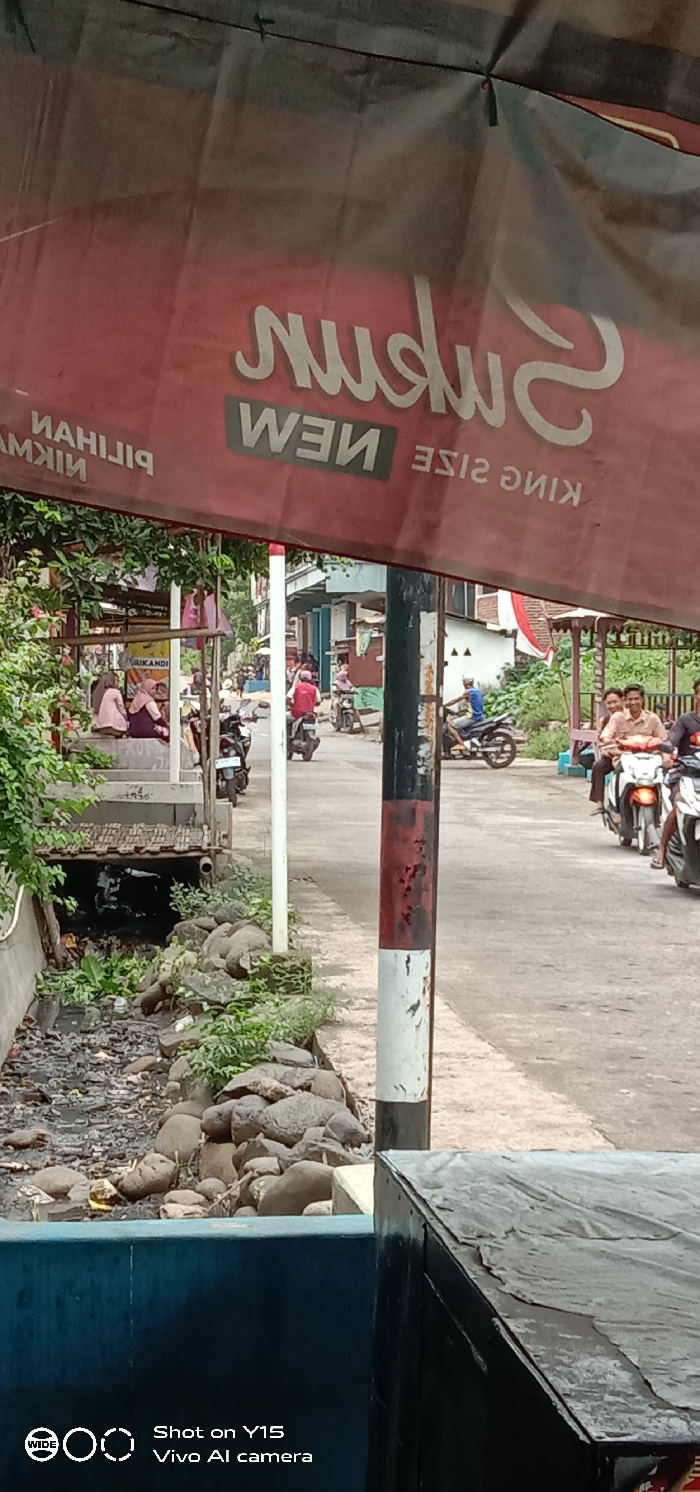 The image captures a candid street scene in Brabe, Indonesia, seemingly taken from a low angle, possibly from beneath a shelter or stall. The foreground is dominated by a red and black banner, partially obscuring the view, with mirrored text that appears to read "SUDOON NEW KING SIZE" (likely a cigarette or product advertisement). Below the banner, to the left, is a drainage ditch or small canal filled with dark water, rocks, and some refuse, bordered by overgrown green plants.
In the midground, a narrow paved road stretches from left to right. Several people are visible. To the left, a group of women, some wearing headscarves, are seated on a concrete structure next to the ditch, appearing to be relaxing or socializing. On the road, multiple individuals are riding or waiting on motorcycles. One man on a motorcycle is smiling at the camera. Another motorcycle is parked near the seated women. A vertical pole, painted in sections of black, white, and red, stands prominently in the center-right, further segmenting the view.
The background reveals more of the local environment, with simple residential or commercial buildings along the roadside and some sparse greenery. The overall impression is that of a typical, active day in a semi-urban or rural Indonesian setting, with daily life unfolding. The sky is not visible, but the lighting suggests it is daytime. The bottom left corner includes a watermark: "Shot on Y15 Vivo AI camera."