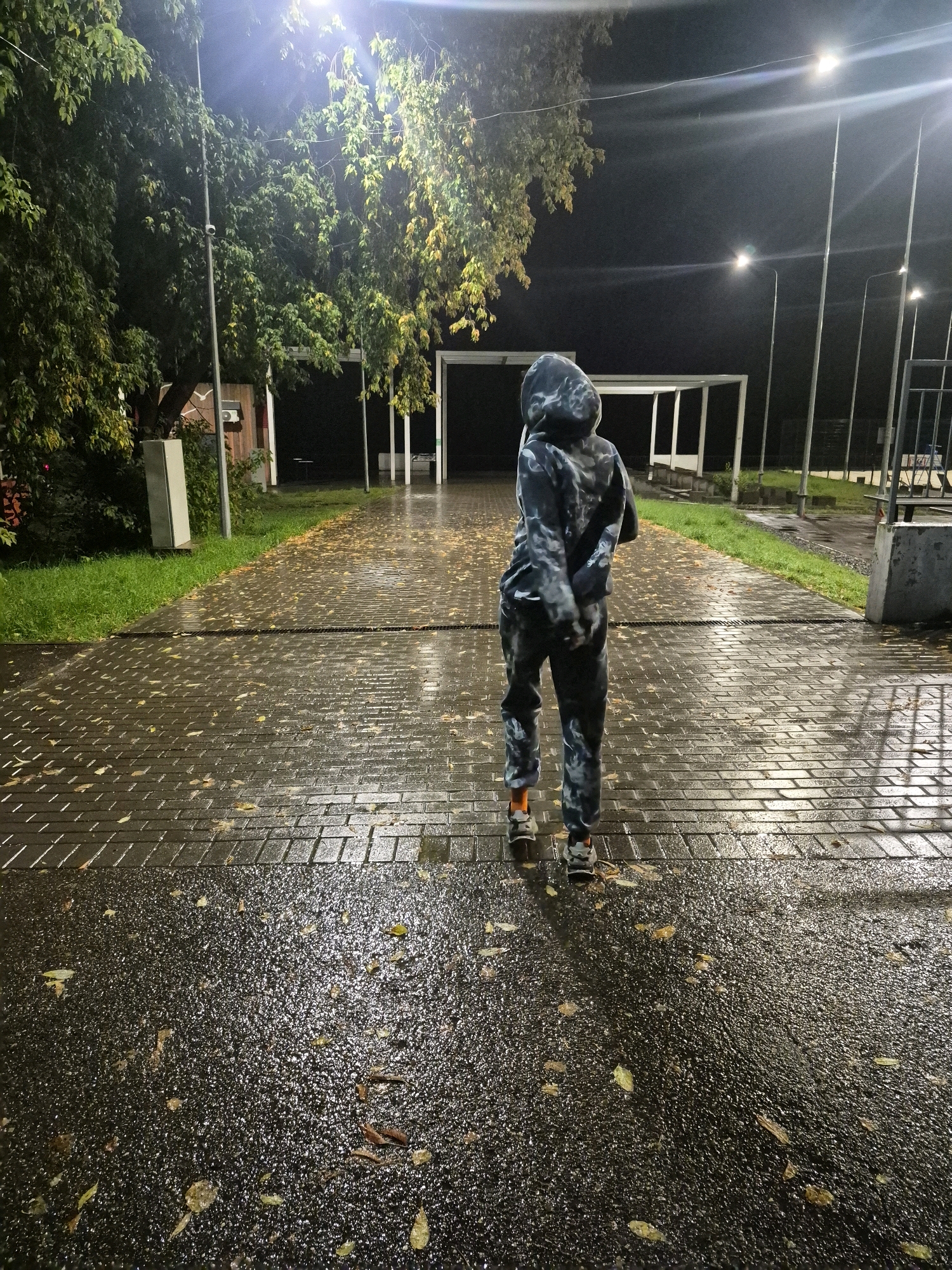 The image depicts a person in a camouflage hooded tracksuit standing on a wet, brick-paved walkway at night. The walkway is illuminated by streetlights, and fallen leaves are scattered across it. To the left of the walkway is a patch of grass, and beyond that, trees with some yellowing leaves. In the background, there are white structures that appear to be part of a covered area or shelter, and further back, more streetlights and fencing. The scene suggests an outdoor area, possibly a park or public space, in Chaikovsky, Russia. The overall impression is one of a damp, dimly lit evening environment.