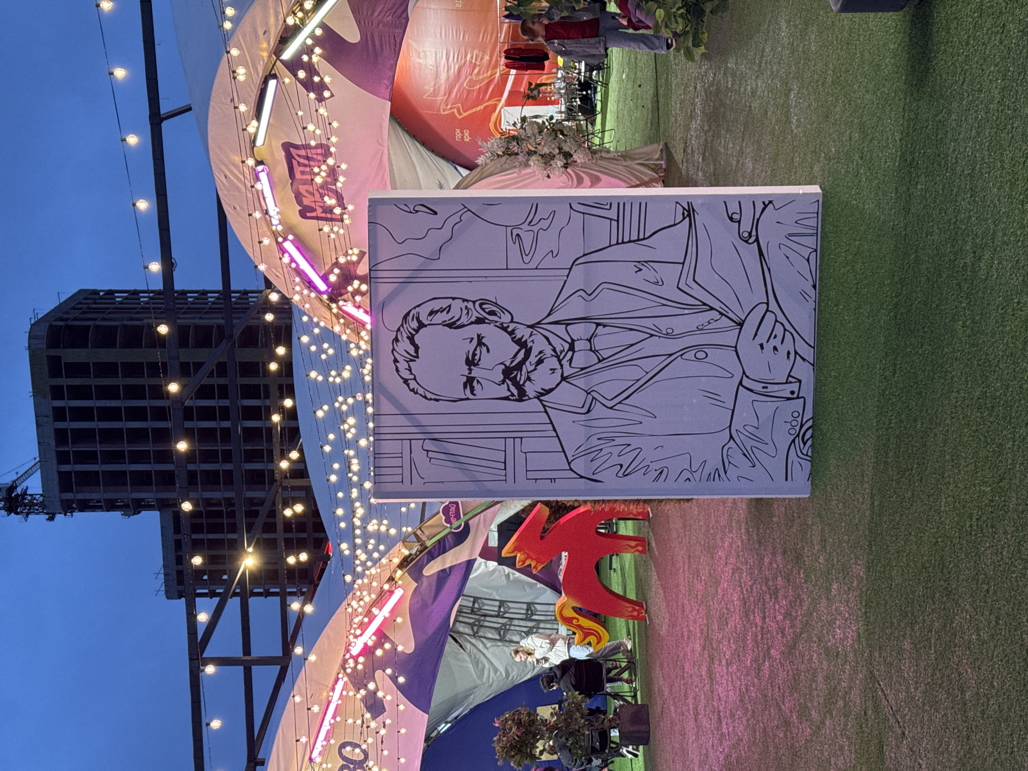 The image captures an outdoor evening scene, likely a public event or marketplace, with a focus on a large black and white drawing of a man's bust. The man in the drawing has a beard and is wearing a suit and tie. The drawing is placed in the foreground, angled so that the man's face is prominent.
Behind the drawing, a large, white, tent-like structure is visible, adorned with strings of warm white lights and accents of pink neon lighting. The structure has a curved shape and appears to be part of a larger setup. In the background, a dark, unfinished building with a visible crane suggests an urban environment, possibly under construction.
The ground is covered with artificial turf, with areas illuminated by pinkish light. Several people are scattered around the area, some sitting on chairs, others standing. One person is visible in the background, near some clothing racks. A bright red, stylized dragon-like sculpture with yellow accents is also present.
The sky is a deep blue, indicating it's dusk or early evening. The overall atmosphere appears festive and lively, with the abundance of lights creating a warm and inviting ambiance. The presence of the drawing, sculpture, and stalls suggests a cultural event or a market. The location context is Izhevsk, Russia. No discernible text is visible on the drawing itself, though there might be some on banners or signs obscured by the lighting and angles.