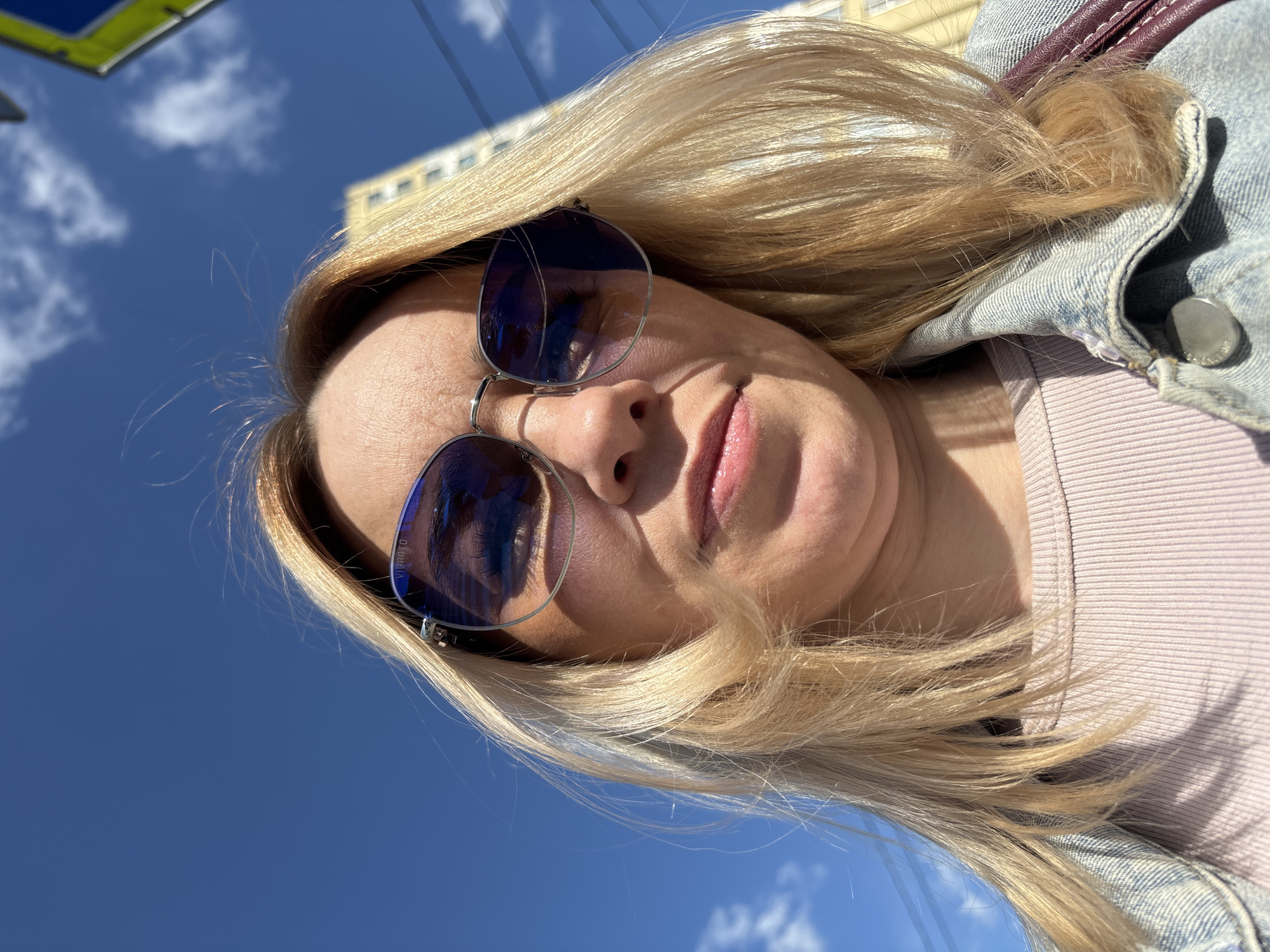 A low-angle selfie captures the upper body of a person, likely female, with long, blonde hair falling around their face and shoulders. The person is looking down towards the camera, with their head slightly tilted. They wear large, roundish, blue-tinted sunglasses that cover their eyes, and the brand "VIBRANTO" is visible on the right lens. Their lips are slightly parted, showing a light pink color.
The individual is dressed in a light pink or lavender ribbed top, layered under a light blue denim jacket featuring a prominent metal button on the front. A dark red or burgundy strap, possibly from a bag, is visible behind the jacket on the upper right side.
The background features a bright, clear blue sky with scattered white clouds, indicating a sunny daytime setting. Strong sunlight creates noticeable shadows on the person's face and clothing. Partially visible in the background are elements of an urban environment: a light yellow building with dark windows behind the person's head, and a green and yellow structure, possibly a sign or part of a building, in the upper left corner. The image depicts a casual outdoor moment.
