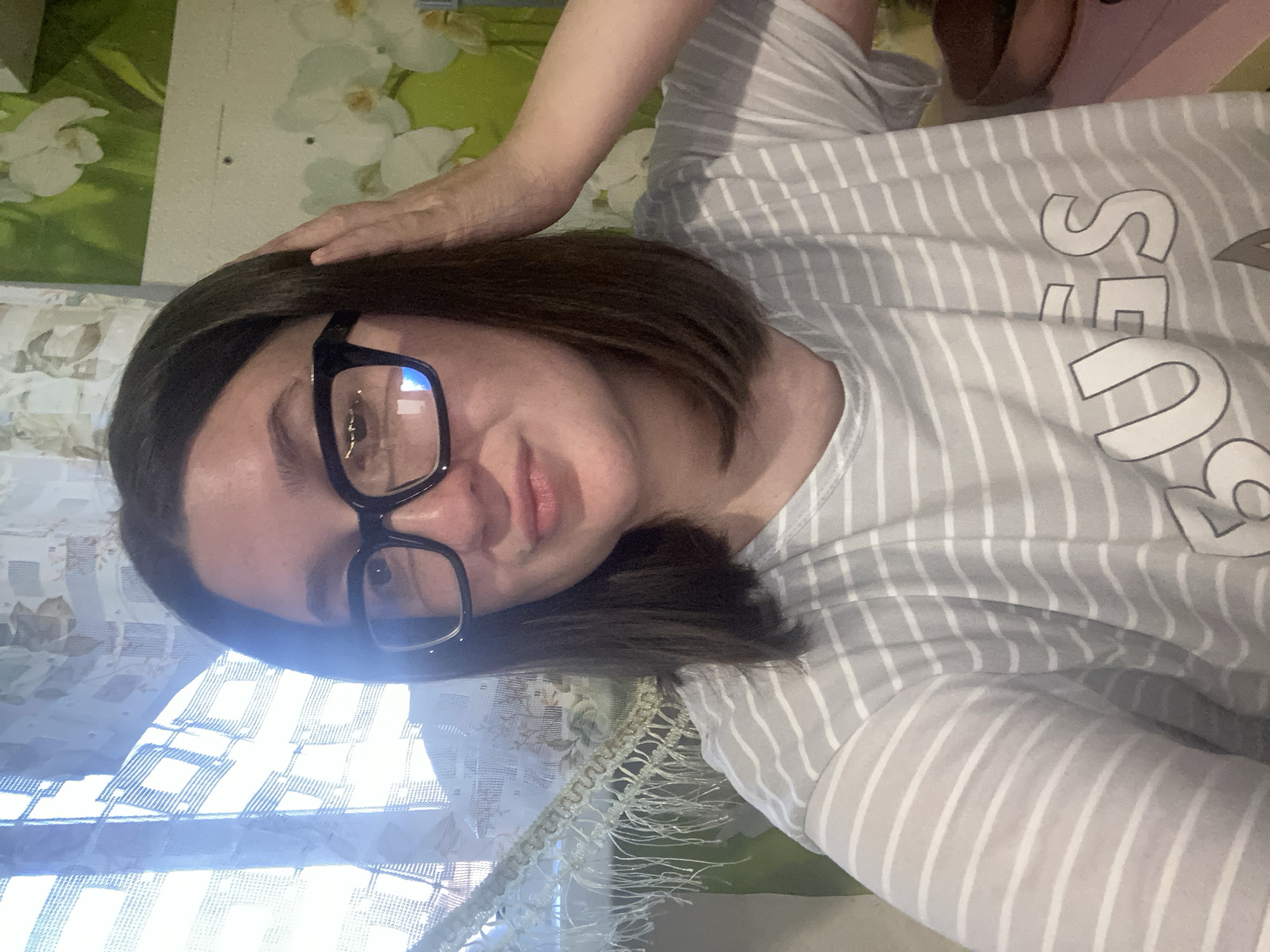 A selfie of a woman with short brown hair and glasses. She is wearing a gray and white striped shirt with white lettering. Her right hand is on her head. Behind her, there is a patterned wall and a curtained window. The location is purportedly Chaikovsky, Russia.