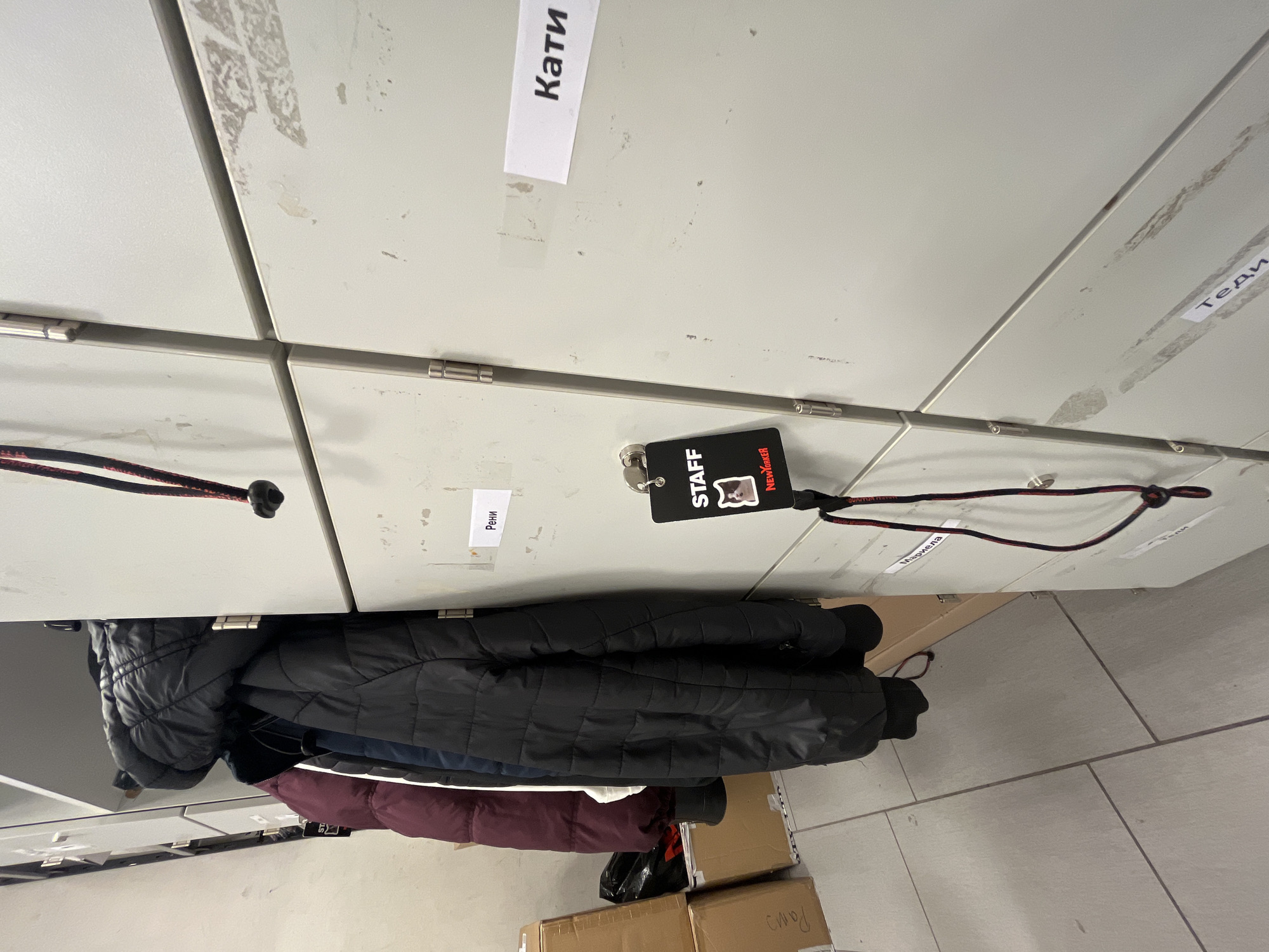 The image displays an interior scene of a staff changing area, likely located within a New Yorker retail store in Sofia, Bulgaria. The primary focus is on a bank of light grey metal lockers, arranged in a grid and showing signs of wear, including scuff marks and adhesive residue. Several locker doors feature white rectangular labels with names written in Cyrillic script, specifically "Рени" (Reni), "Кати" (Kati), "Мариела" (Mariela), and "Теди" (Tedi).
A black "STAFF" identification badge is prominent in the mid-right foreground, hanging from a silver key attached to a red and black braided lanyard. The badge prominently displays a white graphic resembling a cat's head and the "New Yorker" logo in red.
To the left of the lockers, a row of three padded winter jackets hangs on a rack: a dark maroon jacket, a dark blue jacket, and a black jacket. Below these garments and to the far left, several brown cardboard boxes are stacked on a light grey tiled floor. One box partially visible contains a black plastic bag. The walls are light-colored, and the overall environment appears utilitarian.