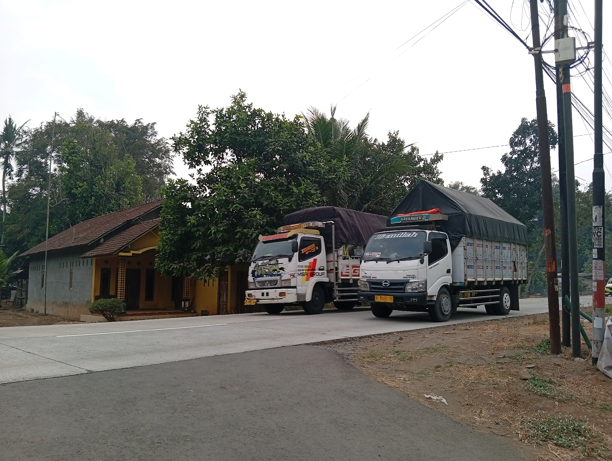 Two trucks are driving on a paved road. The truck on the left is white with red and yellow accents. It has "SAHARA GROUP" written on the side. The truck on the right is also white and has "Bismillah" written on the front. The trucks are driving past a yellow and gray house with a red tile roof. There are trees and utility poles on the right side of the road. The sky is overcast. The scene appears to be in a rural or semi-rural area of Plempukankembaran, Indonesia.