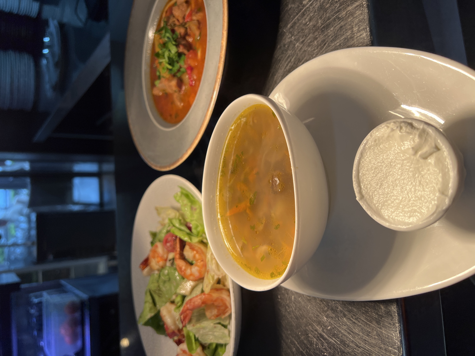 The image shows a table setting with three plates of food. From left to right: a salad with shrimp, a bowl of soup with a side of sour cream, and another bowl of soup. The food appears to be set up in a restaurant. The background is blurred, but there appear to be windows in the upper left side. Based on the lighting and the fact that there is a window, the restaurant may be set outside. However, there is no real information about the precise location to determine it as being in Budva, Montenegro.
