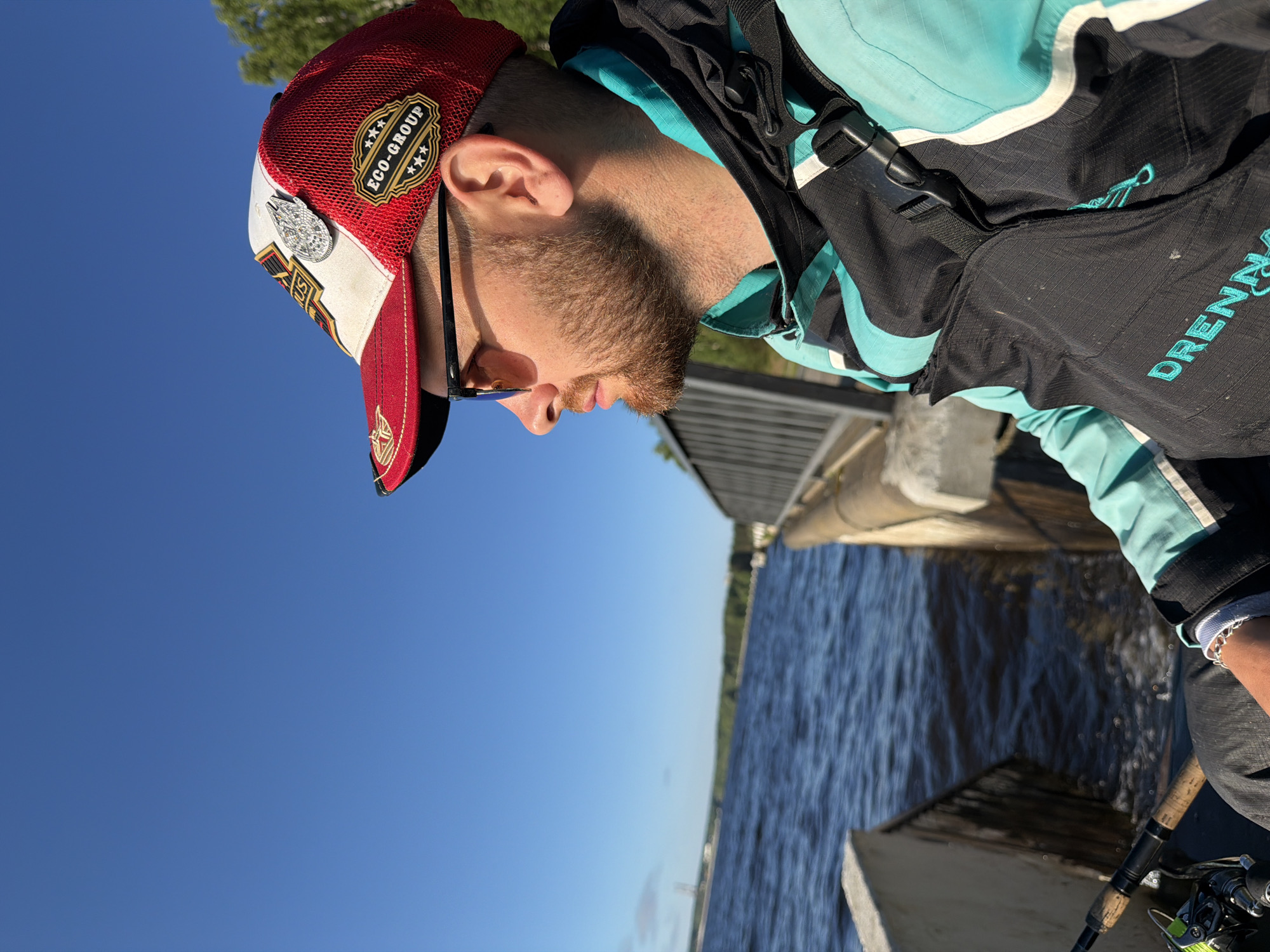 A man with a beard and sunglasses, wearing a red and white baseball cap and a turquoise fishing vest over a jacket, looks out over a body of water. A fishing rod is visible in the foreground. The scene appears to be outdoors, possibly at a dam or embankment, with a clear blue sky and trees in the background. The specific location in Chaikovsky, Russia, cannot be determined from the image alone, but the context suggests a recreational area near water.