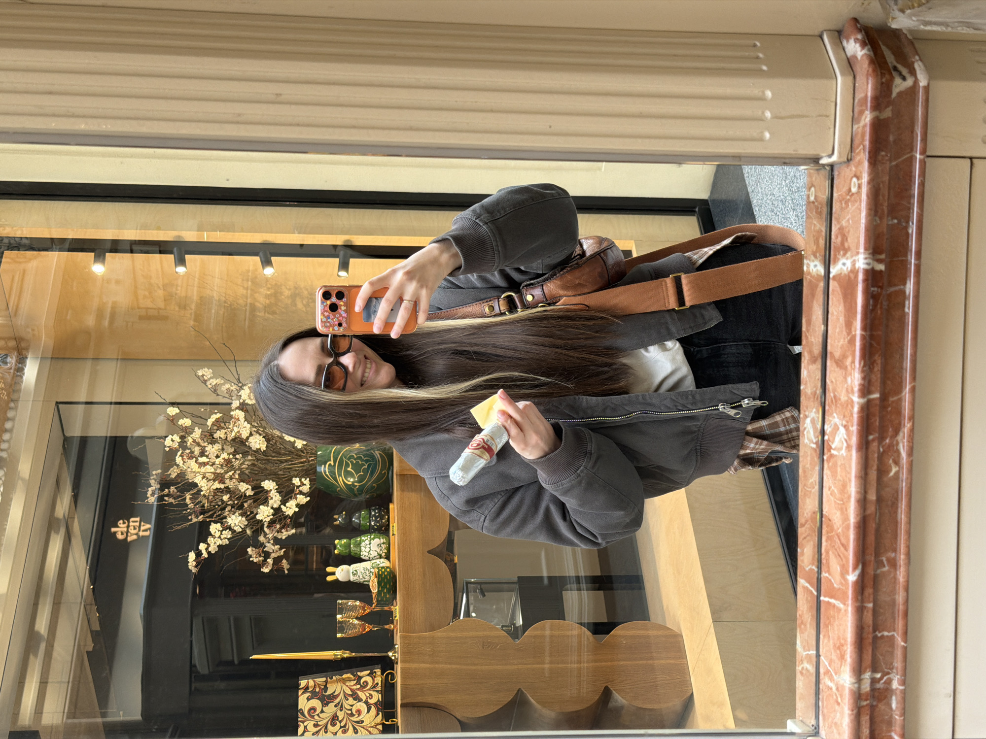 A young woman is standing in front of a store window in Moscow, Russia, taking a selfie. The store window display contains a small bouquet of dried flowers, green glass objects, and wooden shelves with decorated notebooks, glasses, and small animal figurines. She wears a dark gray jacket, black pants, and a tan shoulder bag. The frame around the window is made of marble. She is holding a packaged food product in one hand and a cell phone in the other, which she uses to take the photo.