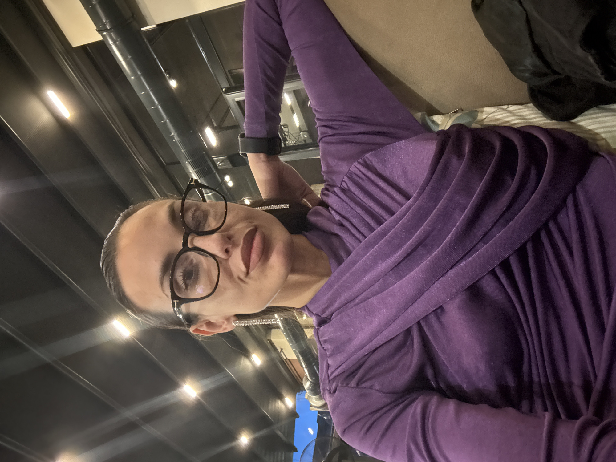 A woman wearing glasses and a purple shirt is lying on a couch indoors. The background shows a modern interior with visible ductwork and lighting fixtures. No specific details indicate the location is Pleven, Bulgaria.