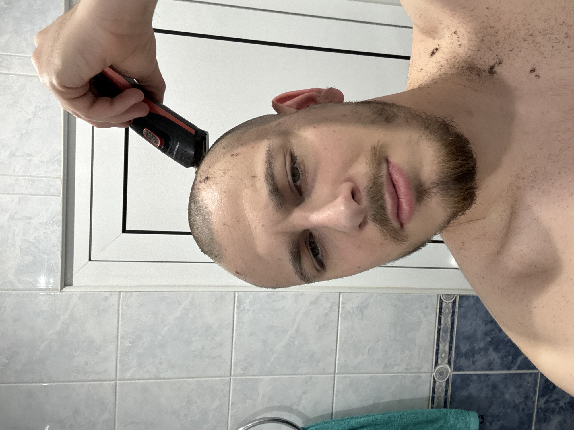 A man is in a bathroom, shaving his head with an electric razor. The bathroom has light blue and white tiled walls and a white-framed window. A rolled-up green towel is visible at the bottom of the image. No specific location in Stara Zagora, Bulgaria can be determined from the image.