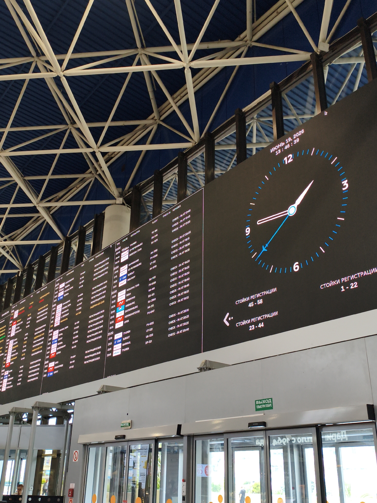 This is an image of the interior of an airport terminal, likely in Sochi, Russia, given the text "SOCHI" on the departure board. The scene is dominated by a large, digital departure board displaying flight information, including flight numbers, destinations, and check-in times. The board is illuminated and shows a wide array of text in Russian. To the right of the departure board, a large digital clock displays the current time as 13:46:39 on June 19, 2026. The clock face is stylized, with a plane icon as the second hand. Below the clock, text indicates "CHECK-IN COUNTERS" with corresponding numbers.
The ceiling of the terminal features an intricate white metal truss structure supporting a dark blue roof. The lighting suggests it is daytime, though the interior is somewhat dimly lit, creating a mood typical of an airport environment.
In the foreground, large glass automatic doors are visible, with a green exit sign above them that reads "EXIT ONLY IN CASE OF FIRE". Reflections on the glass suggest the outside is bright, possibly sunny. A few people are partially visible near the doors, including one person in uniform at a podium on the left. The overall impression is that of a busy, modern airport.
