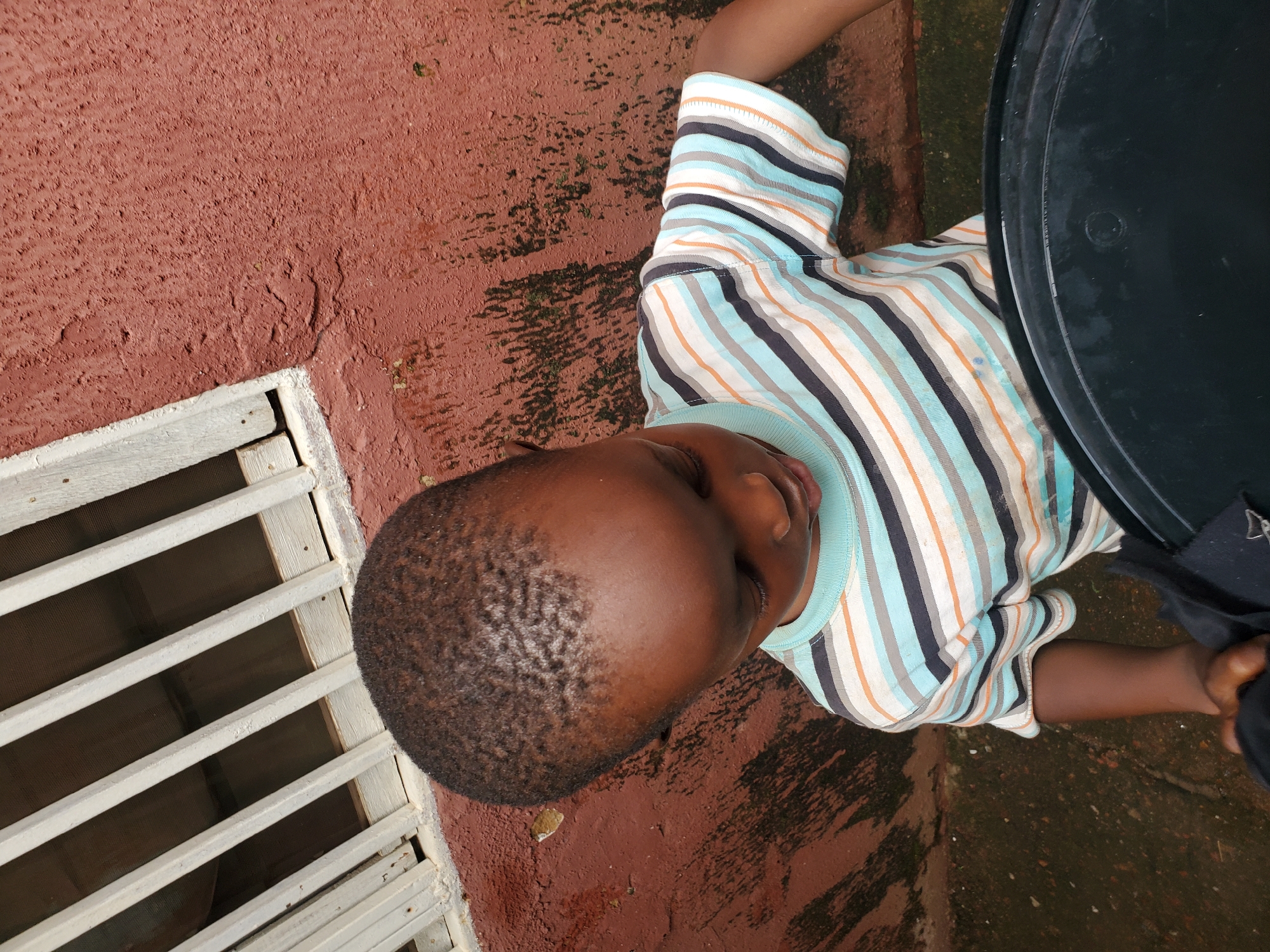 A young child lies on a reddish-brown, textured surface that appears to be pavement or concrete. The child is wearing a horizontally striped t-shirt with colors of blue, white, orange, and grey. The child's head is tilted, and their eyes are closed or looking down, giving an impression of rest or sleep.
To the left of the child, there is a white, slatted structure, possibly a window grate or a vent, with a dark opening behind it. To the right of the child, a large, dark, circular object is partially visible, with a textured surface. The ground around the child is wet in some areas, suggesting recent rain or a damp environment. There are also patches of green moss or algae on the ground, particularly near the slatted structure and extending towards the right side of the frame.
The overall lighting suggests it is daytime, but the exact time is not clear. The scene appears to be outdoors, possibly in an urban or residential area given the pavement and grate. There is no visible text in the image. The child's expression is neutral, making it difficult to determine any specific emotions. The setting implies a casual or domestic environment in Jos, Nigeria.