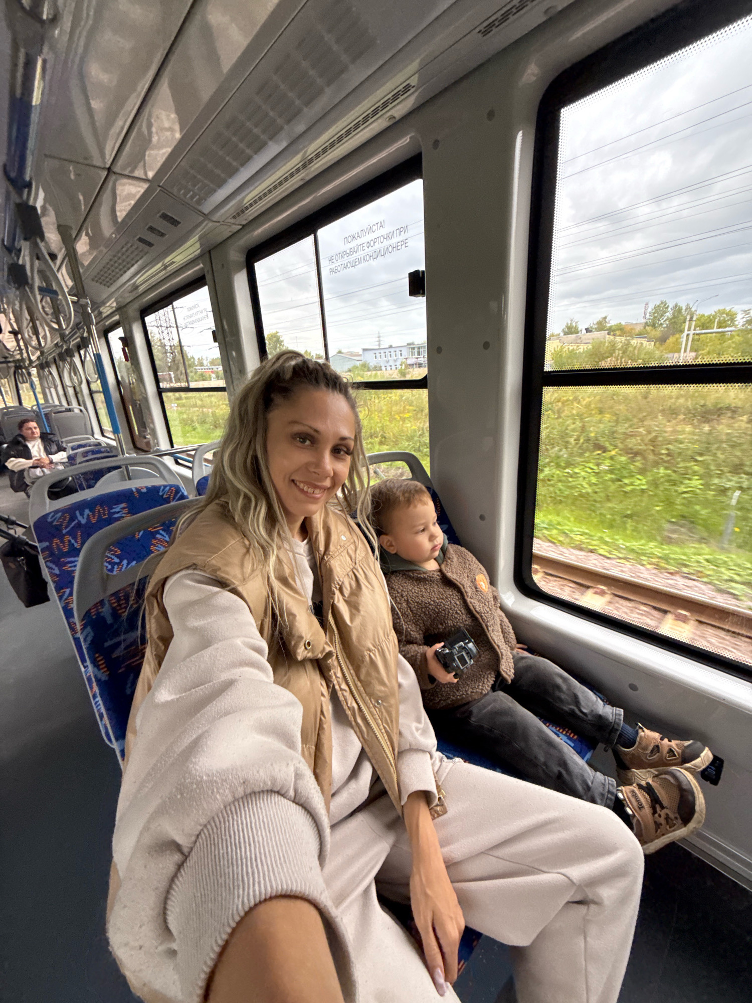 A woman and a young child are seated on a train in Rusanovo, Russia. The woman is in the foreground, smiling at the camera and wearing a tan puffer vest over a cream-colored tracksuit. Her hair is light brown and pulled back in a high ponytail. The child is seated next to her, facing away from the camera and looking out the window. He is wearing a brown, textured jacket and dark pants, and holding a black camera. The train interior features blue and white patterned seats, and large windows that show a green, grassy landscape and railway tracks outside. The sky is overcast. Another passenger is visible further down the aisle on the left.