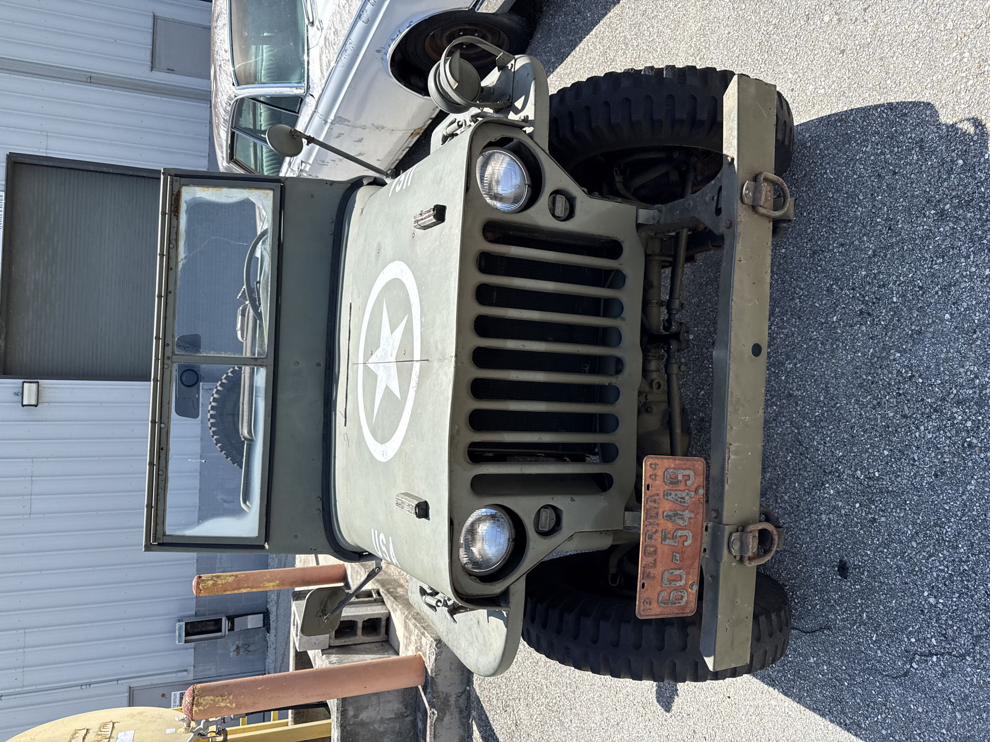 This is a front-facing, medium-shot photograph of a vintage olive drab military jeep parked outdoors on an asphalt surface in Indiantown, Florida, USA. The jeep is the primary subject, positioned centrally in the frame. It features a classic grille with vertical slats, round headlights, and a white star insignia on the hood. A cracked windshield is visible, and a spare tire is mounted behind it. The jeep has a Florida license plate with the number "6D-5449" and the year "44". To the right of the jeep, a weathered, light-colored sedan is partially visible. In the background, a utilitarian building with a corrugated metal exterior and a large rolling door is present. Adjacent to the building, there are several concrete blocks and two cylindrical orange posts, one of which is weathered and peeling. A yellow structure, possibly part of a utility pole, is visible on the far left. The scene is brightly lit by sunlight, casting shadows on the ground.