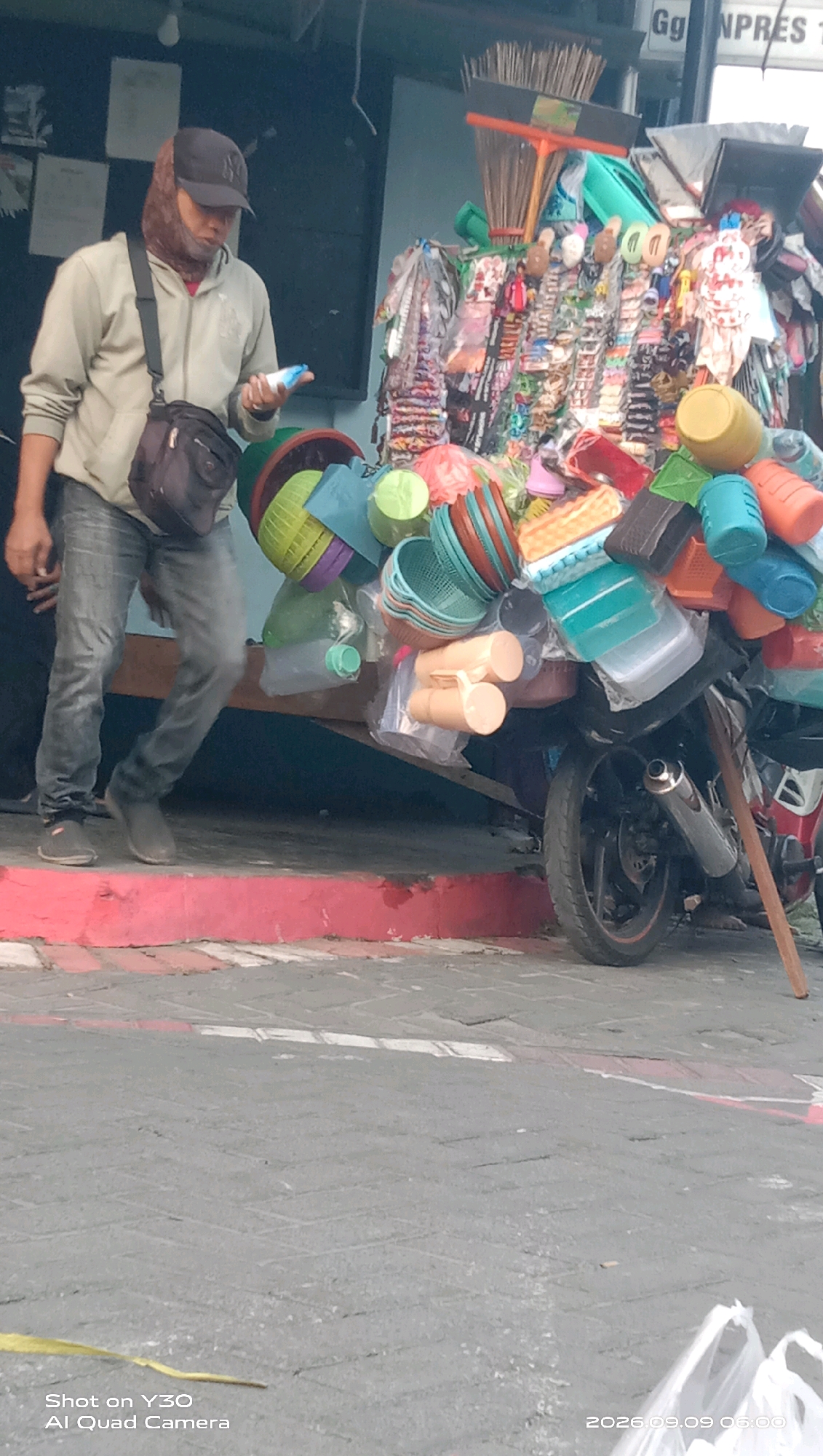 A man wearing a light-colored hooded jacket, a dark baseball cap, and blue jeans stands on a red-painted curb, holding a light blue object in his right hand. He has a dark crossbody bag slung over his left shoulder. Beside him, a motorcycle is loaded with various items, including plastic containers, baskets, brooms, and other merchandise. The motorcycle is propped up by a wooden stick. The scene appears to be outdoors, with a building in the background and a paved ground in the foreground. The image is dated 2026.09.09 06:00.