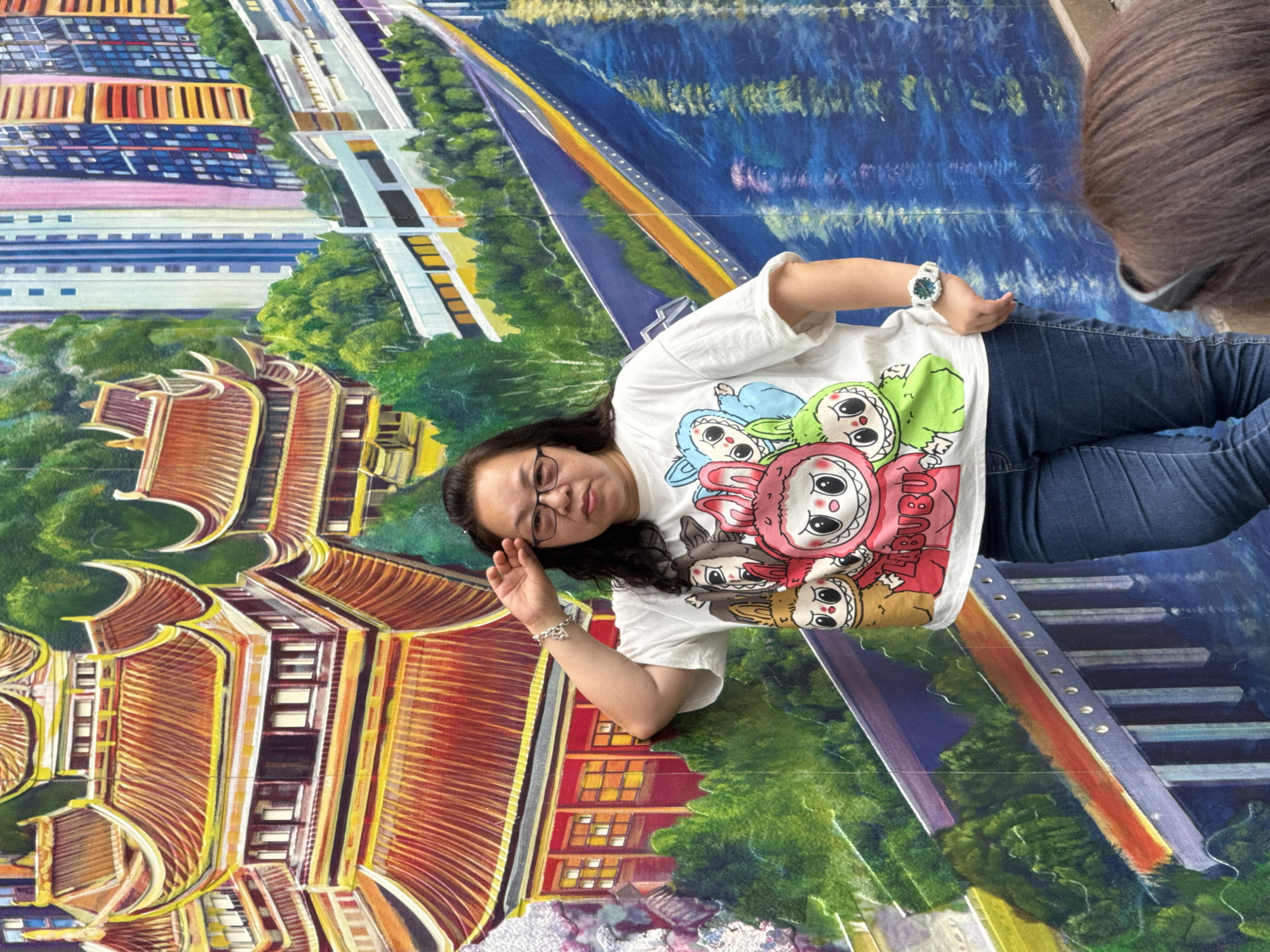 A person is standing in front of a mural. The mural depicts a scene of Yuzhong District, China. The mural includes a traditional Chinese building with a red roof, modern buildings, and a river. The person is wearing glasses, a white t-shirt with cartoon characters, and blue jeans. They are standing with their hand on their glasses and looking towards the camera. The environment is an urban street setting.
