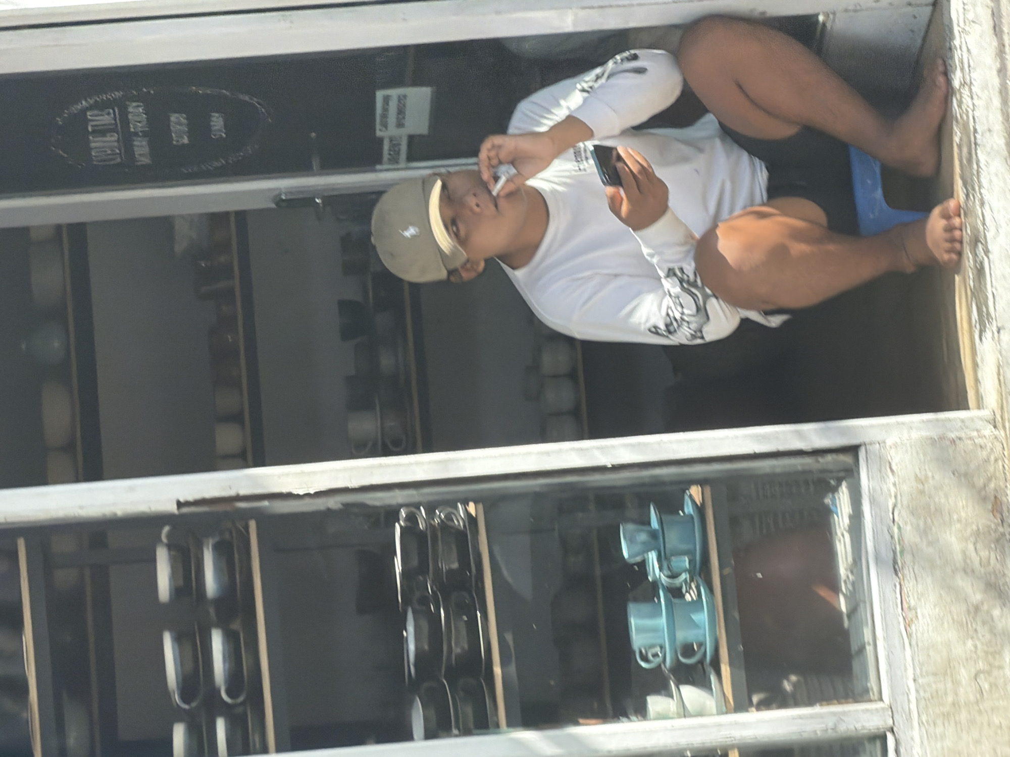 A man is seated in what appears to be a doorway or window opening of a storefront, viewed through a reflective glass pane. He is wearing a beige baseball cap, a white long-sleeved shirt with dark designs on the sleeves, and dark shorts. His feet are bare. He is holding a black smartphone in his left hand and what looks like a cigarette or vape device to his mouth with his right hand, appearing to be taking a puff. He is looking directly forward.
The man is sitting on a small, light blue plastic stool. Behind him, visible through the glass, are shelves lined with various cups or mugs, some dark-colored and others light blue. These items suggest the setting is a cafe or a shop that sells beverages or kitchenware. The overall lighting is bright, indicating daytime.
Through the upper part of the glass, a dark rectangular sign is visible. The legible text on the sign reads: "KEDAI TAO", "MONDAY-FRIDAY", "SATURDAY", and "SUNDAY". Below "SATURDAY" and "SUNDAY," there appear to be additional, less clear markings, possibly indicating operating hours. The context of North Kuta, Indonesia, suggests a tropical or warm climate. The image has a strong reflection on the glass, showing elements of the outside environment.