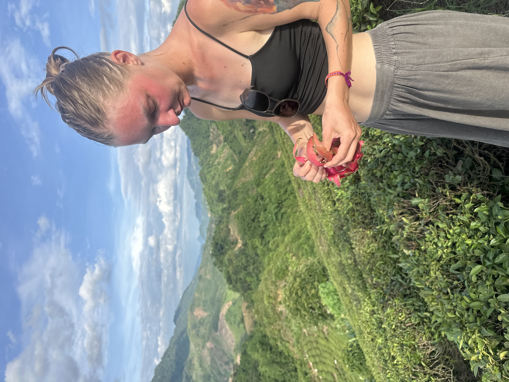 A young woman with blonde hair pulled into a bun stands in front of a scenic mountainous landscape in To Mua, Vietnam, holding a dragon fruit. She is wearing a black spaghetti strap tank top and a long grey skirt. Her face, particularly her cheeks and forehead, is flushed red, suggesting sunburn. Sunglasses are clipped to the front of her tank top. She has a tattoo on her right arm. The background features lush green terraced hills, indicative of tea plantations or agricultural cultivation, under a bright blue sky with scattered white clouds. The terrain is characterized by rolling hills and valleys.