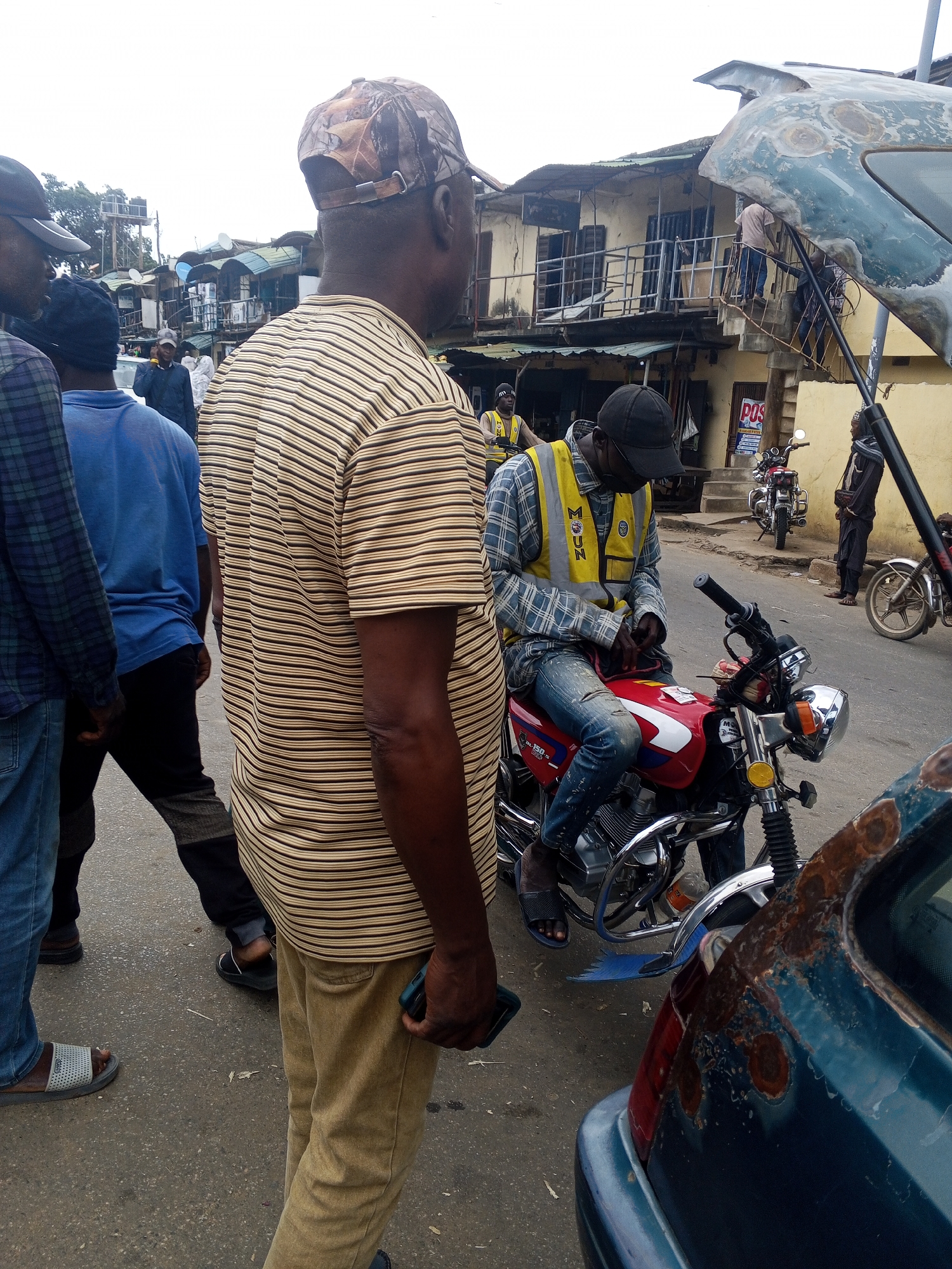 In Okene, Nigeria, a street scene unfolds with several individuals and vehicles. In the foreground, a person in a striped shirt and camouflage cap stands facing away from the camera. To their left, another person wearing a blue shirt walks away. A man wearing a plaid shirt and a yellow vest sits on a red motorcycle, looking down. Further back, a red motorcycle is parked. Buildings with corrugated metal roofs and balconies line the street. Another motorcycle is visible to the right of the frame, parked near a weathered, blue car with visible rust. A person in dark clothing stands near the car. The ground is paved with asphalt. The overall impression is a bustling urban environment with everyday activities taking place.