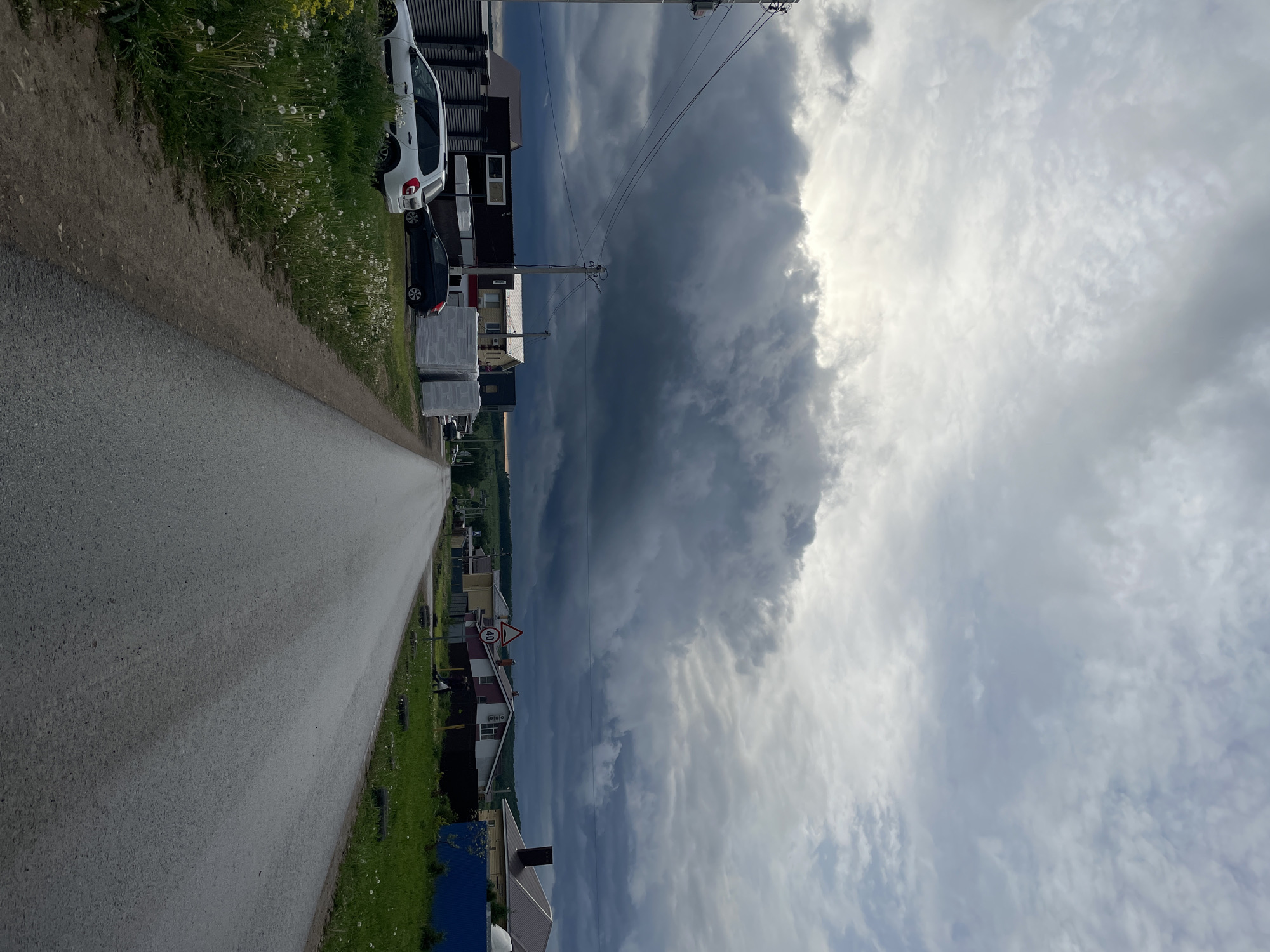 A street in Эстачи, Russia is shown. A gray asphalt road stretches into the distance. The roadside features lush green grass and dandelions. Parked cars, including a white car and a dark car, are visible. Buildings are seen along the road. A utility pole with overhead wires stands tall. The sky is filled with a mixture of dark, ominous storm clouds and bright, sunny clouds.